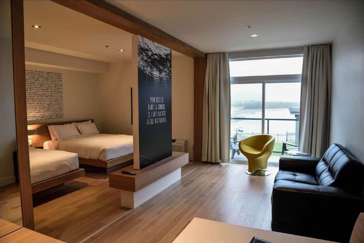 A modern hotel room with a bed, a sofa, a yellow chair, and a large window with a view.