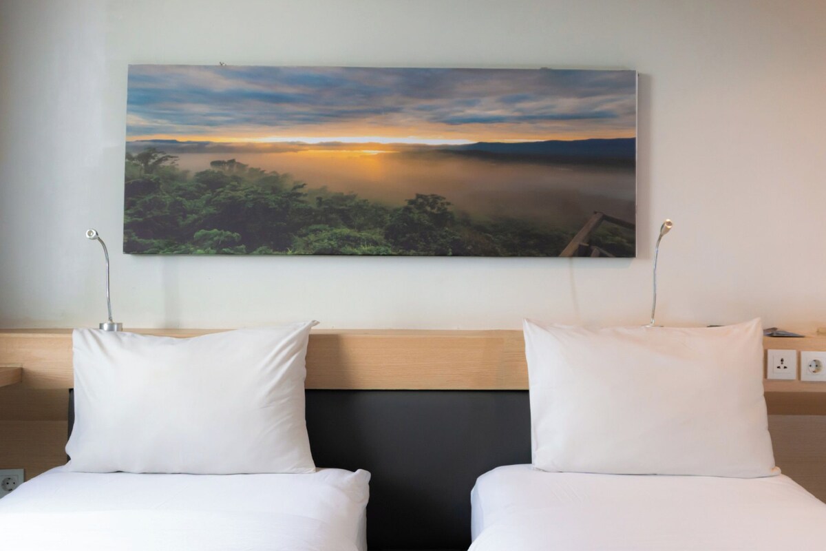 Een hotelkamer met twee enkele bedden, een groot schilderij van een landschap en een houten hoofdeinde.