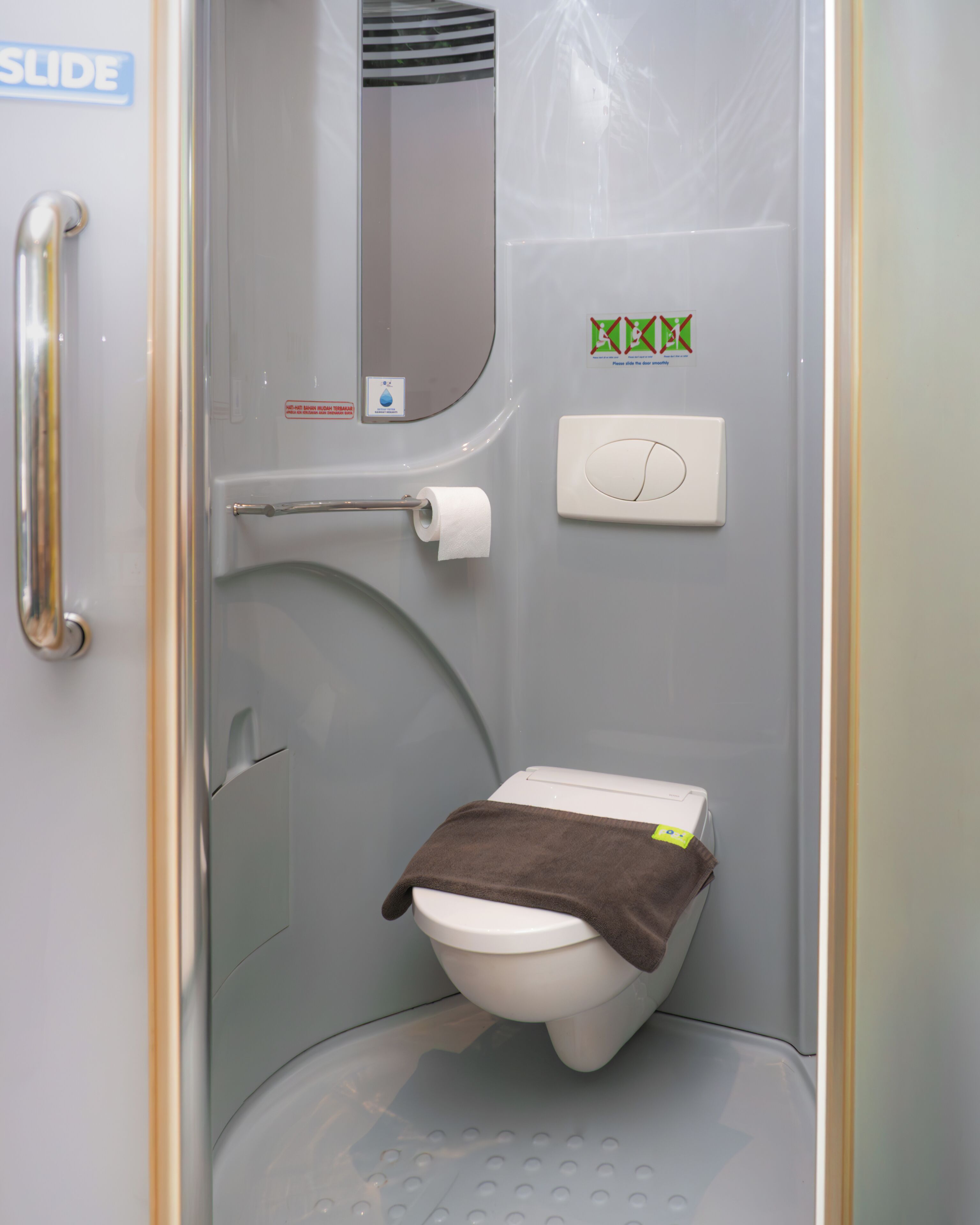 Une petite salle de bain fermée, équipée de toilettes, d’une barre d’appui, d’une serviette et d’un miroir.