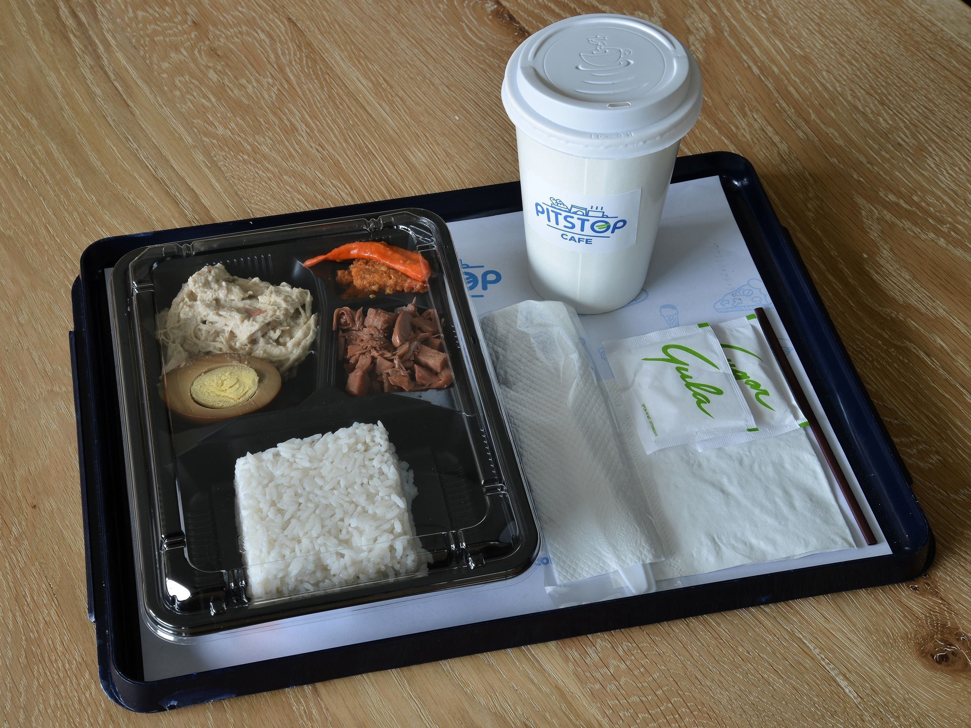 Bandeja de comida con arroz, un huevo duro, carne desmenuzada, un cangrejo y una taza de café sobre una mesa de madera.
