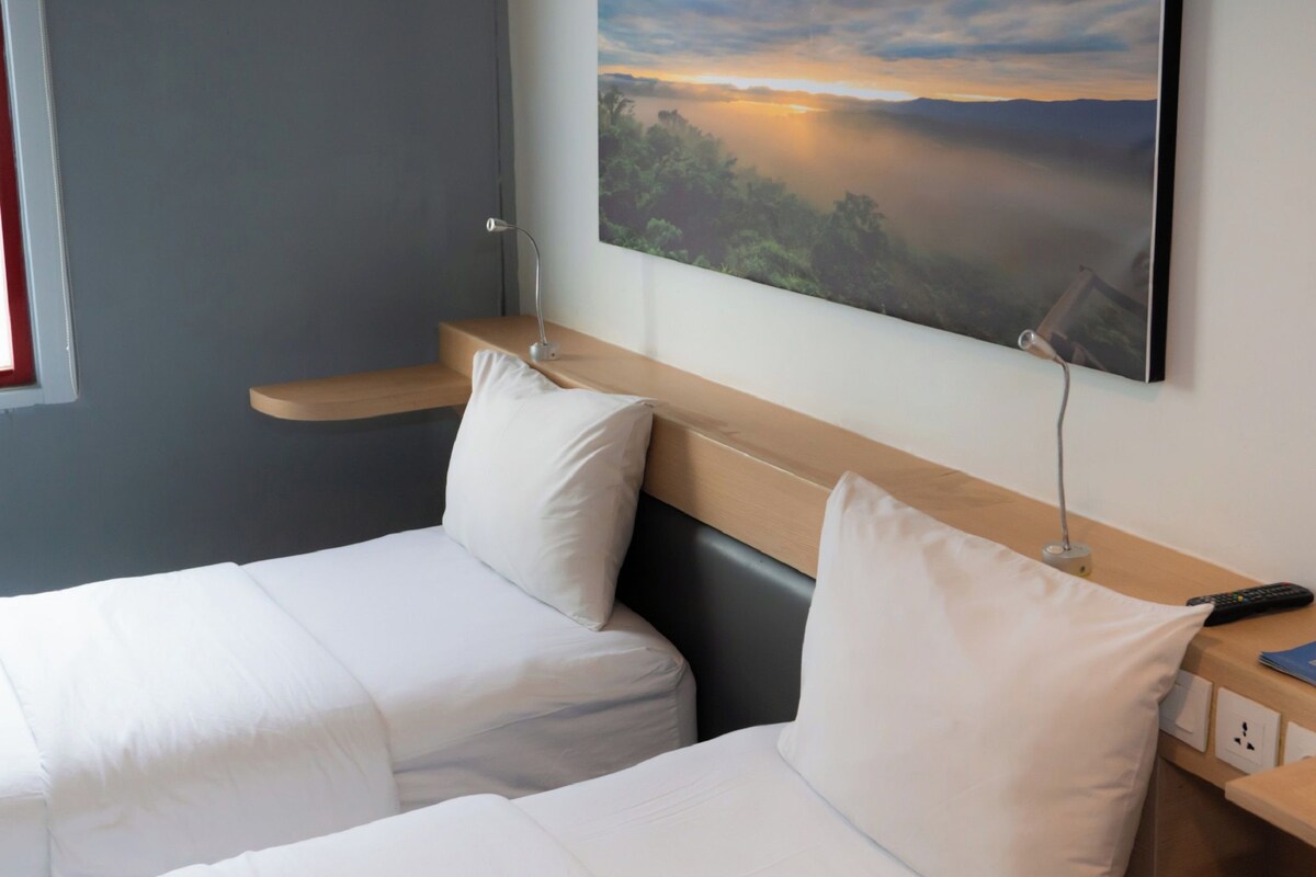 Ein Hotelzimmer mit zwei Betten, einem Landschaftsbild und einem Schreibtisch mit Lampe und Fernbedienung.