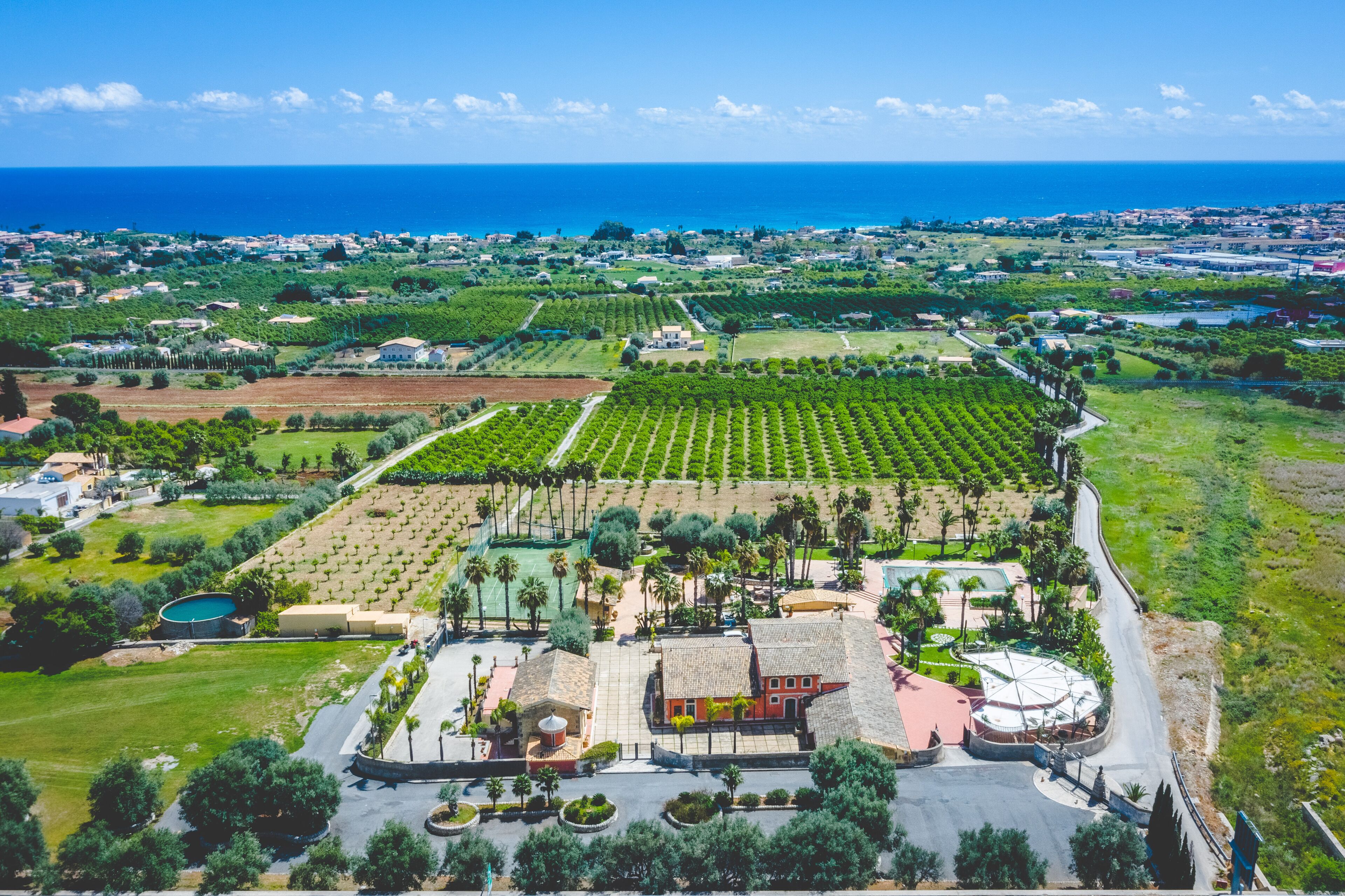 An aerial view of a coastal property with vineyards, a swimming pool, and a large building with a red roof.