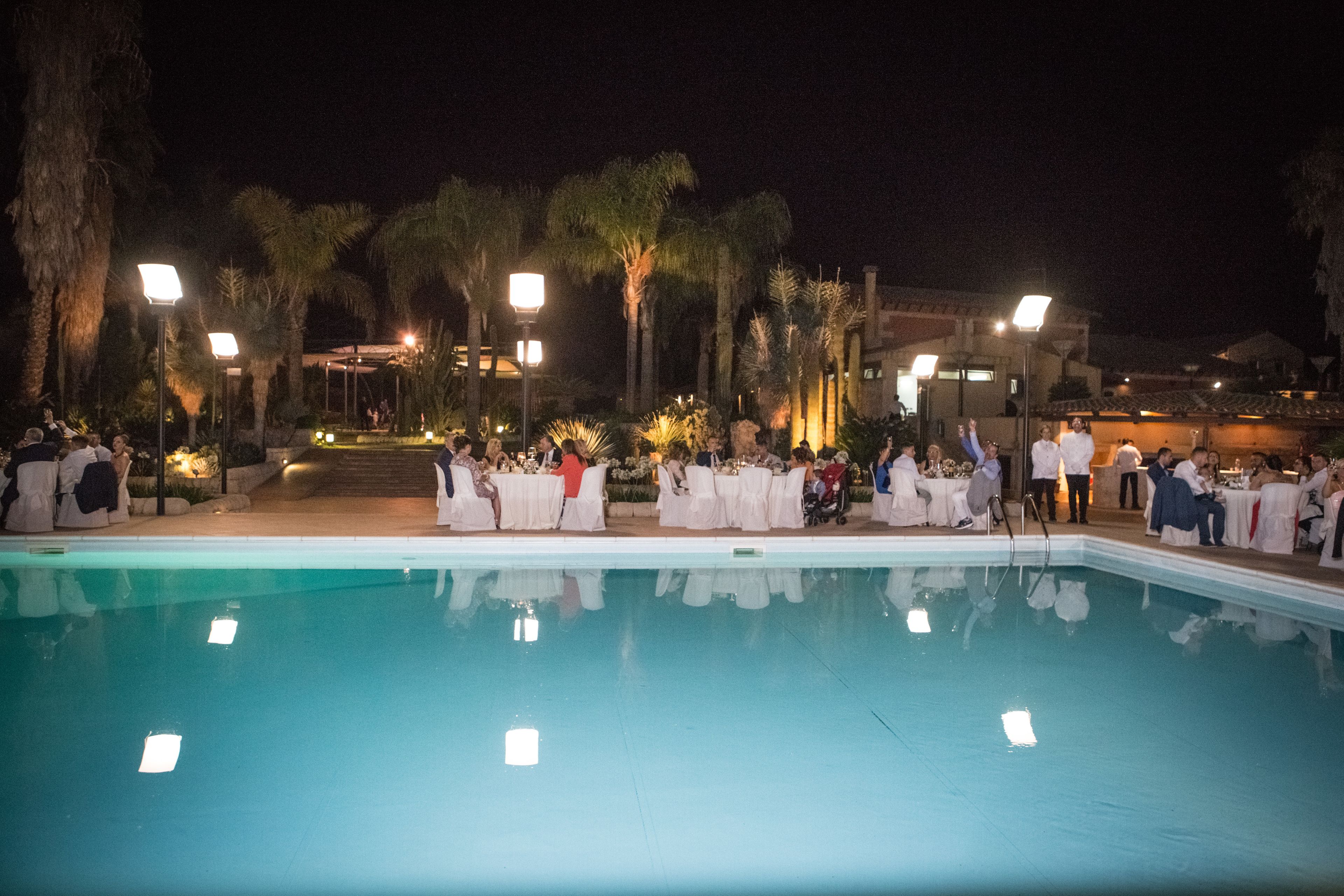 Une réception organisée au bord de la piscine, où les invités sont assis à des tables, entourés de palmiers et éclairés par des lampes suspendues.