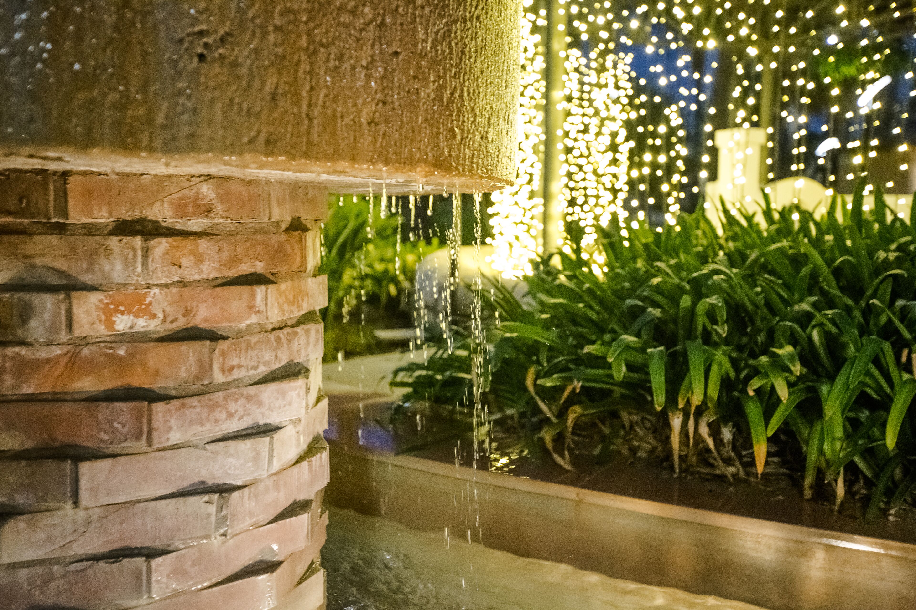 A stone fountain with water flowing into a rectangular basin, surrounded by lush green plants.