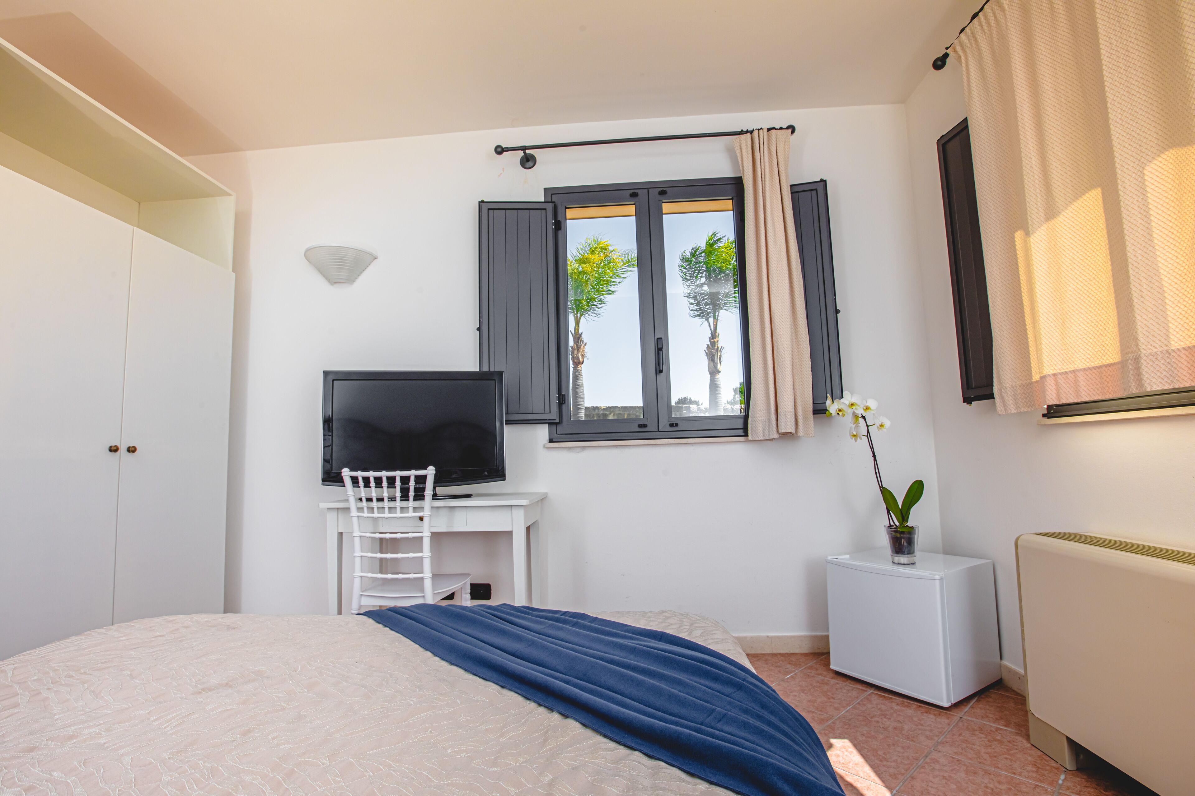 Ein Schlafzimmer mit einem Bett, einem Fernseher, einem Schreibtisch mit Stuhl, einem Fenster mit Vorhängen und einem Heizkörper.