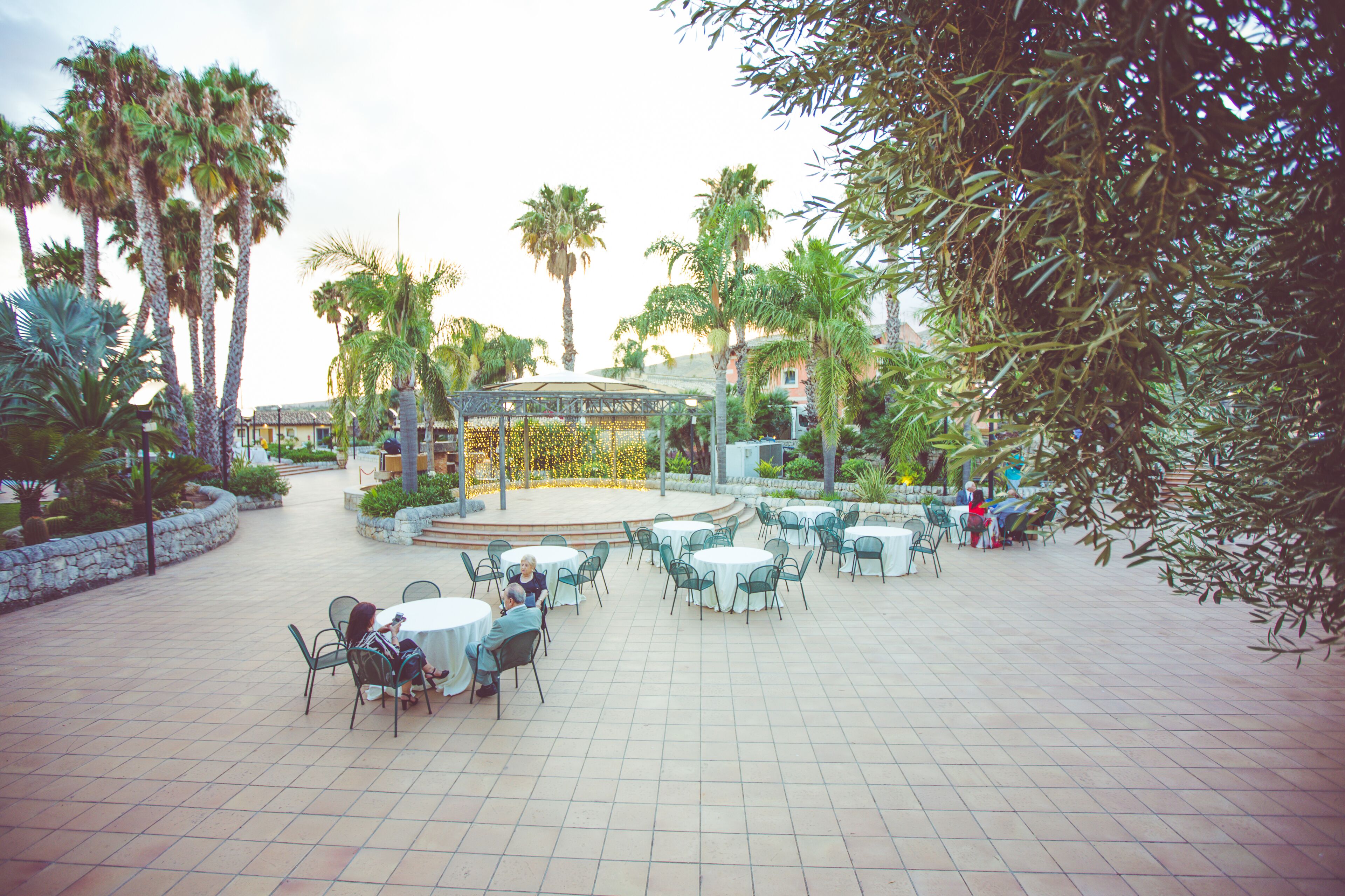 An outdoor patio area with round tables and chairs, surrounded by palm trees and a gazebo.