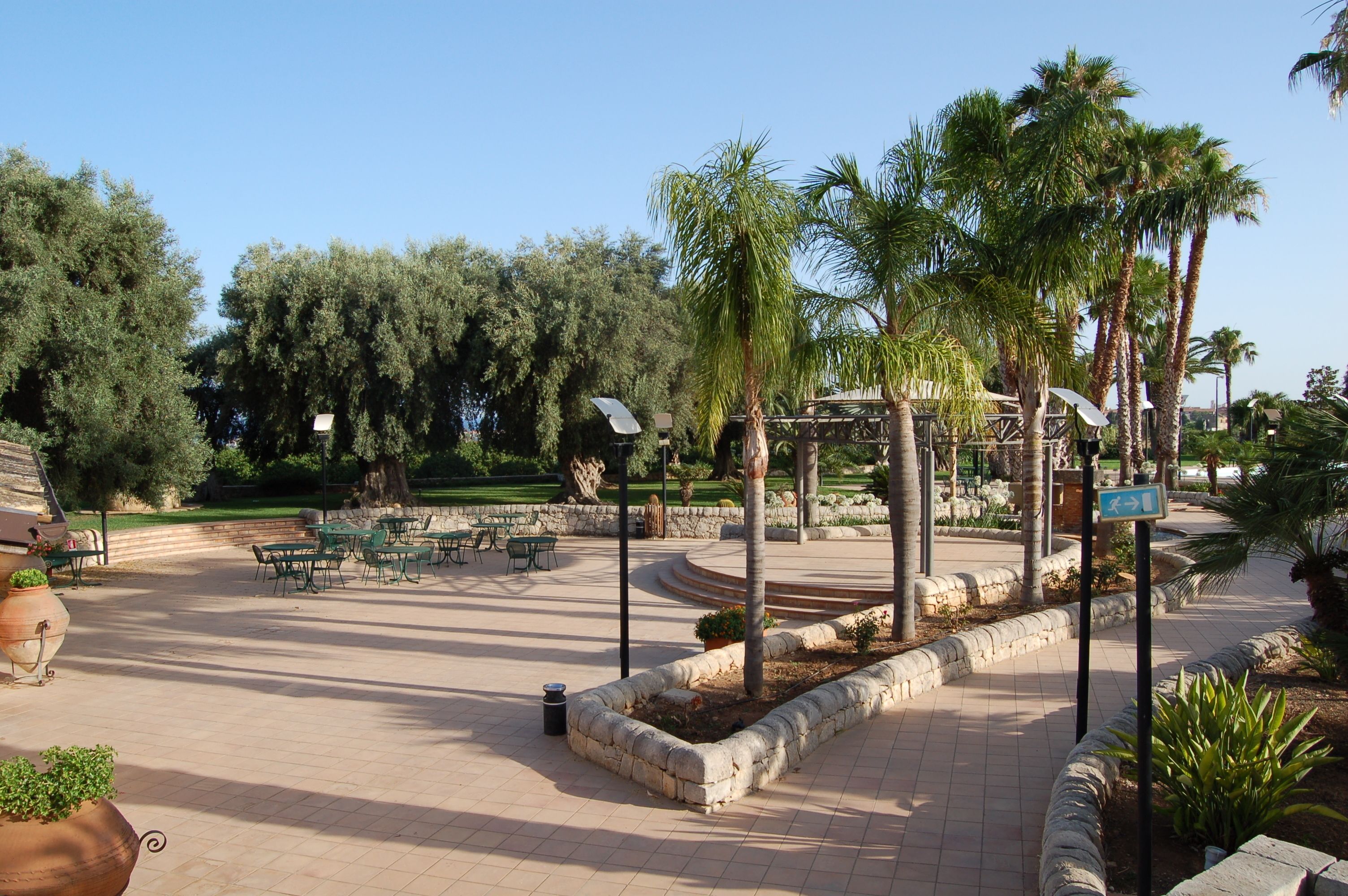 An outdoor patio area with stone flooring, green chairs, and tables, surrounded by palm trees and a stone wall.