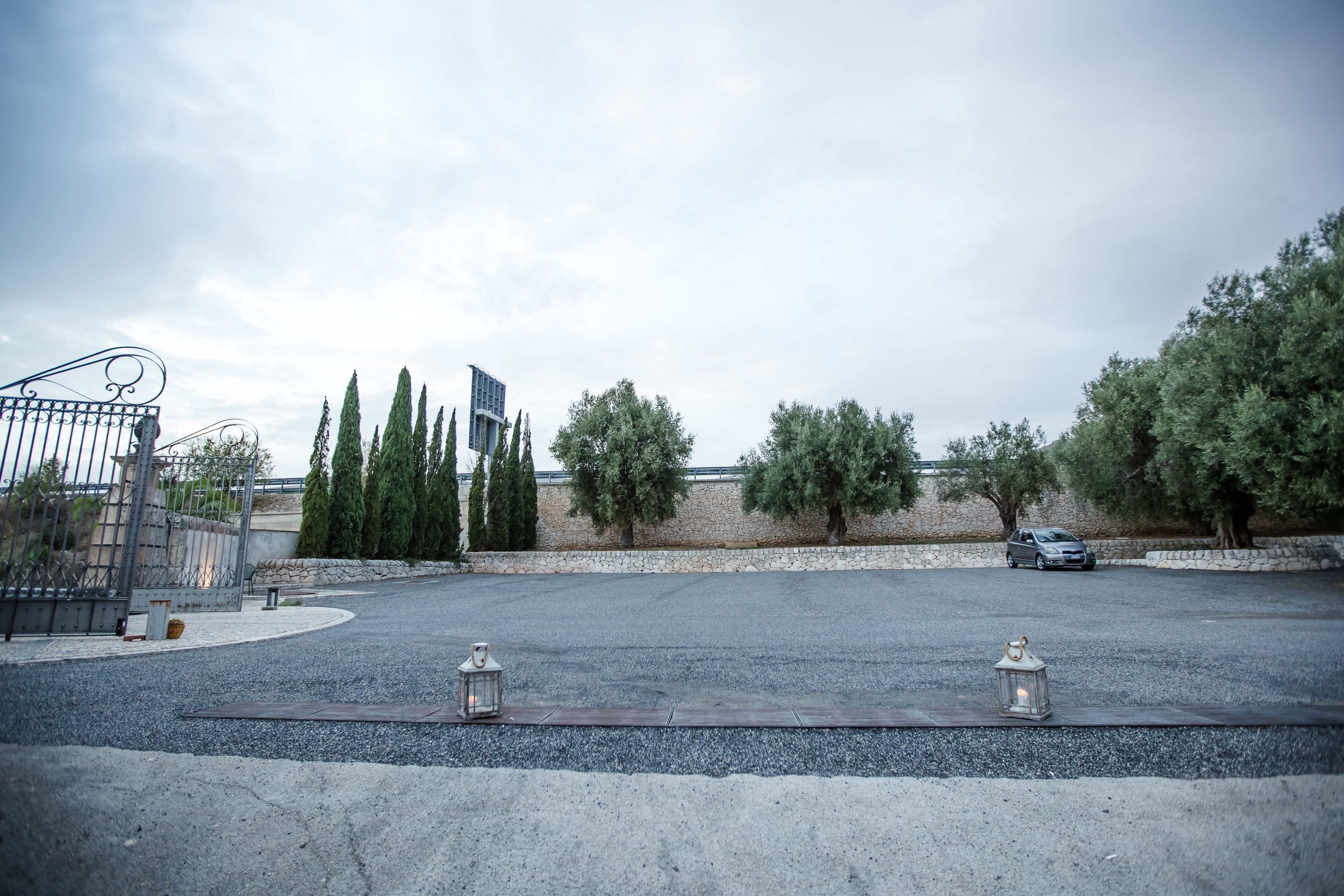 An empty parking lot with a paved road, a metal fence, and a stone wall with trees.