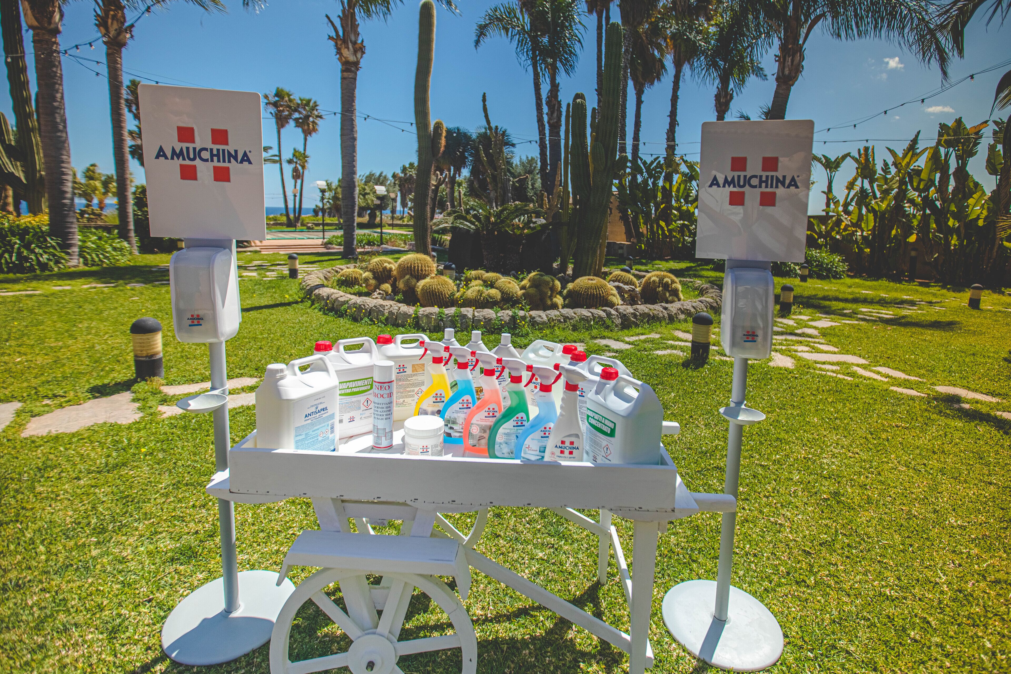 A garden area with a white cart displaying various Amuchina products, surrounded by palm trees and cacti.