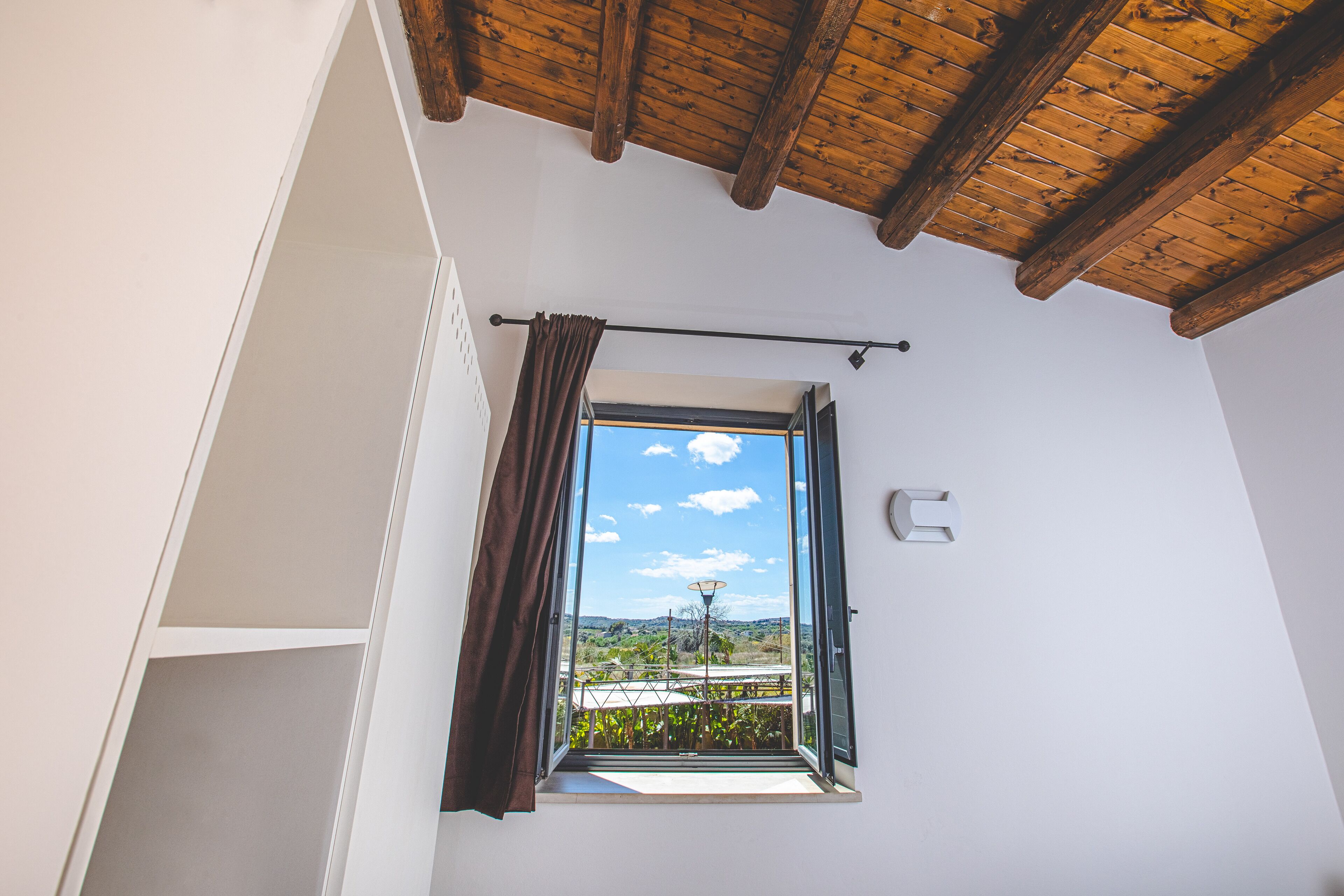 A room with a wooden ceiling, a window with a view of the outdoors, and a white wall with a light switch.