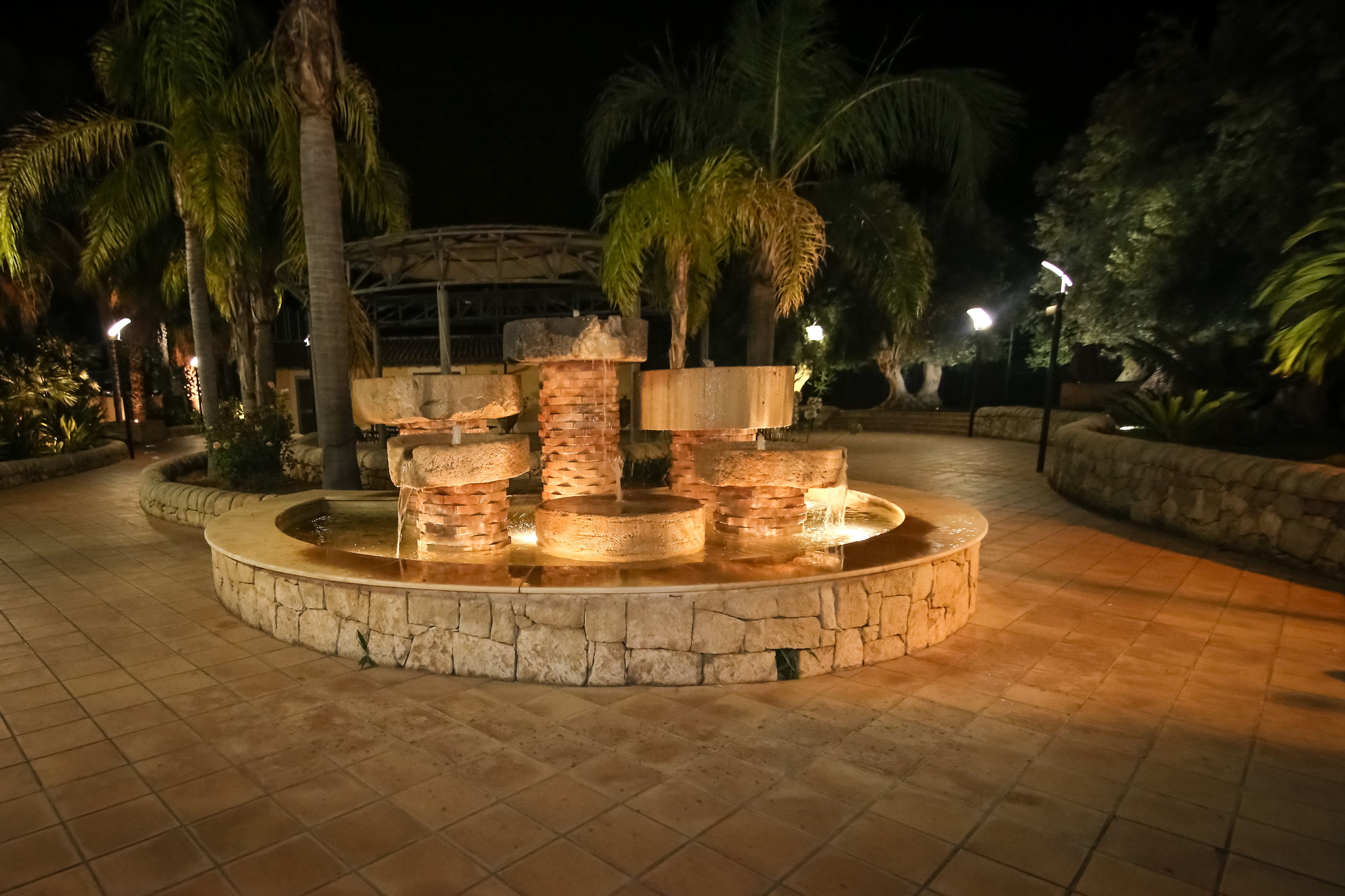 A nighttime outdoor area with a circular stone fountain, palm trees, and stone walls.