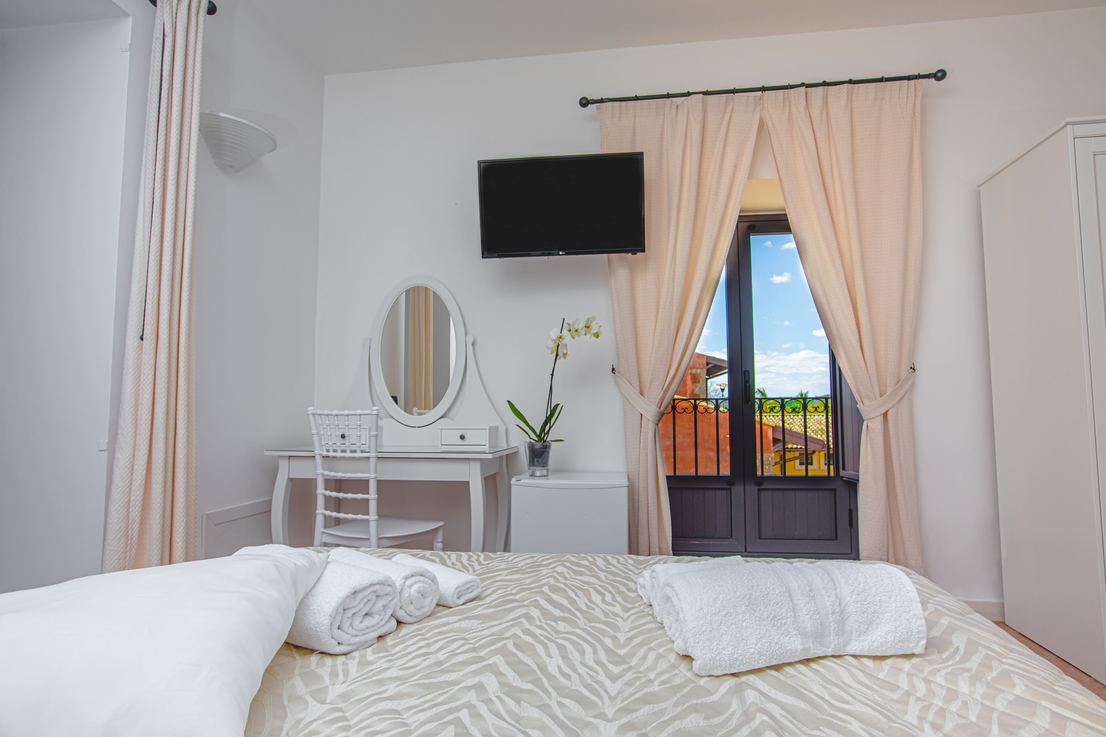 A bedroom with a bed, a desk with a mirror, a TV, and a window with curtains.
