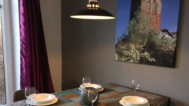 A dining table set with plates, glasses, and cutlery, a hanging light fixture, and a painting of a tower on the wall. - Ferienwohnung Zacharias (Bad Hersfeld)