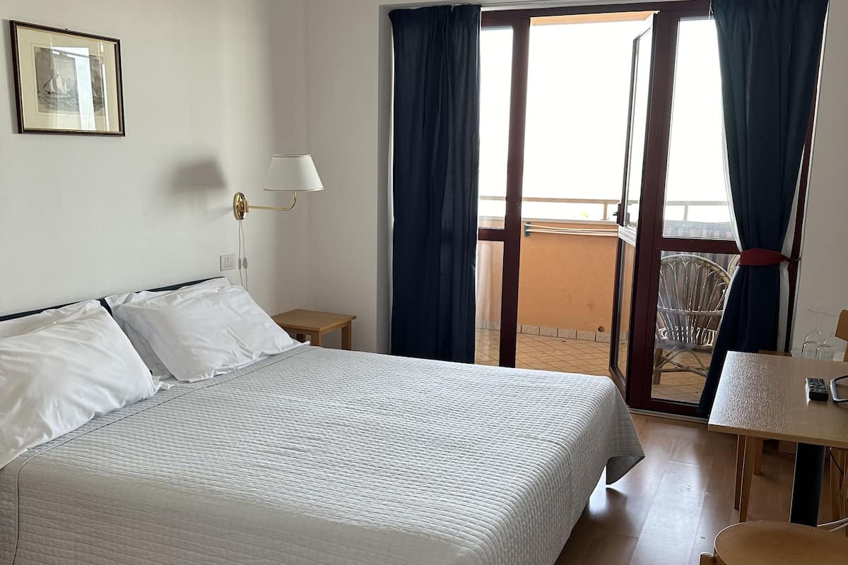 Une chambre d’hôtel avec un grand lit, une table de chevet avec une lampe, une petite table avec une chaise et un balcon avec des rideaux.