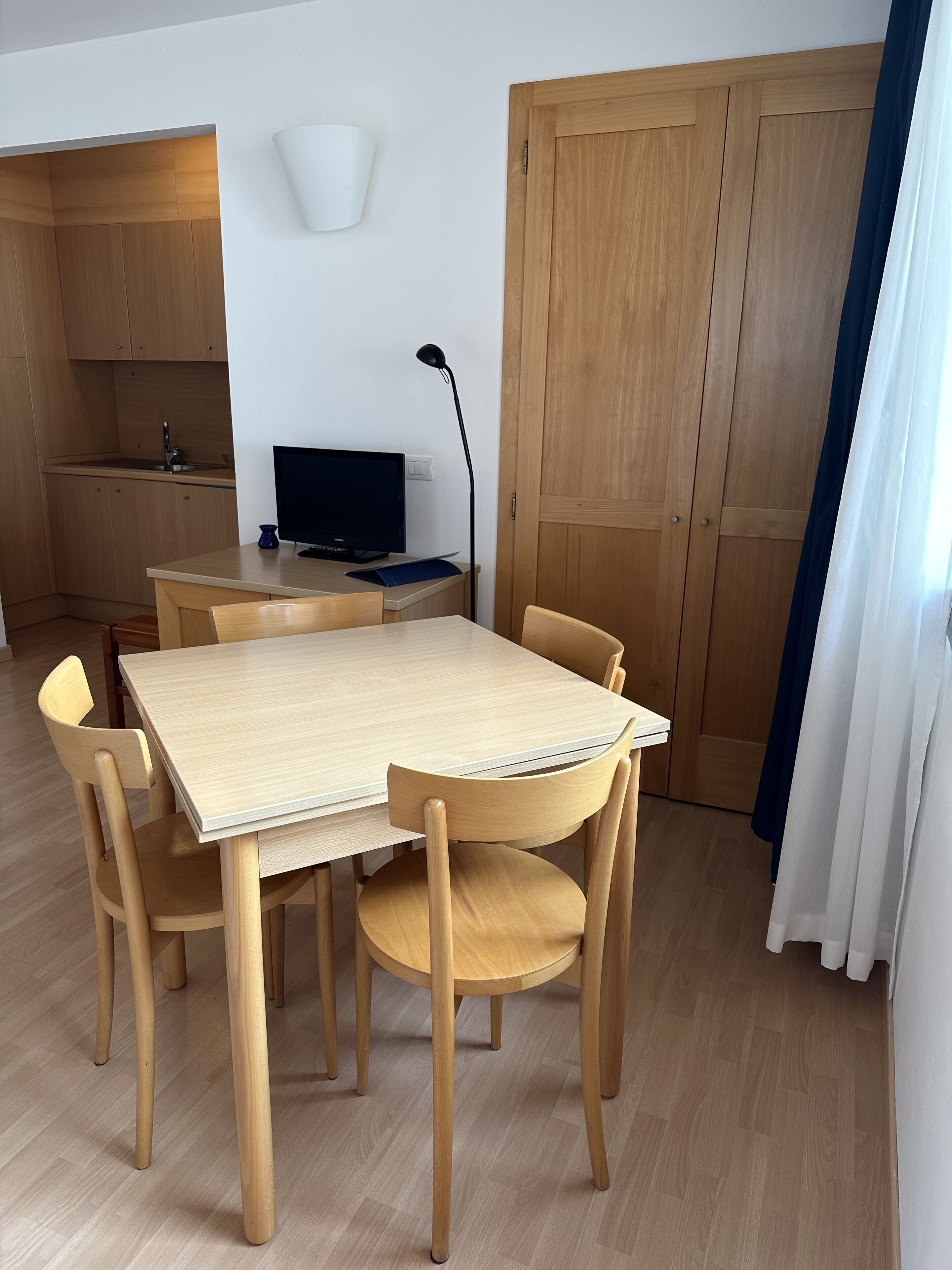 A modern kitchen with wooden cabinets, a dining table with chairs, a TV, and a lamp.