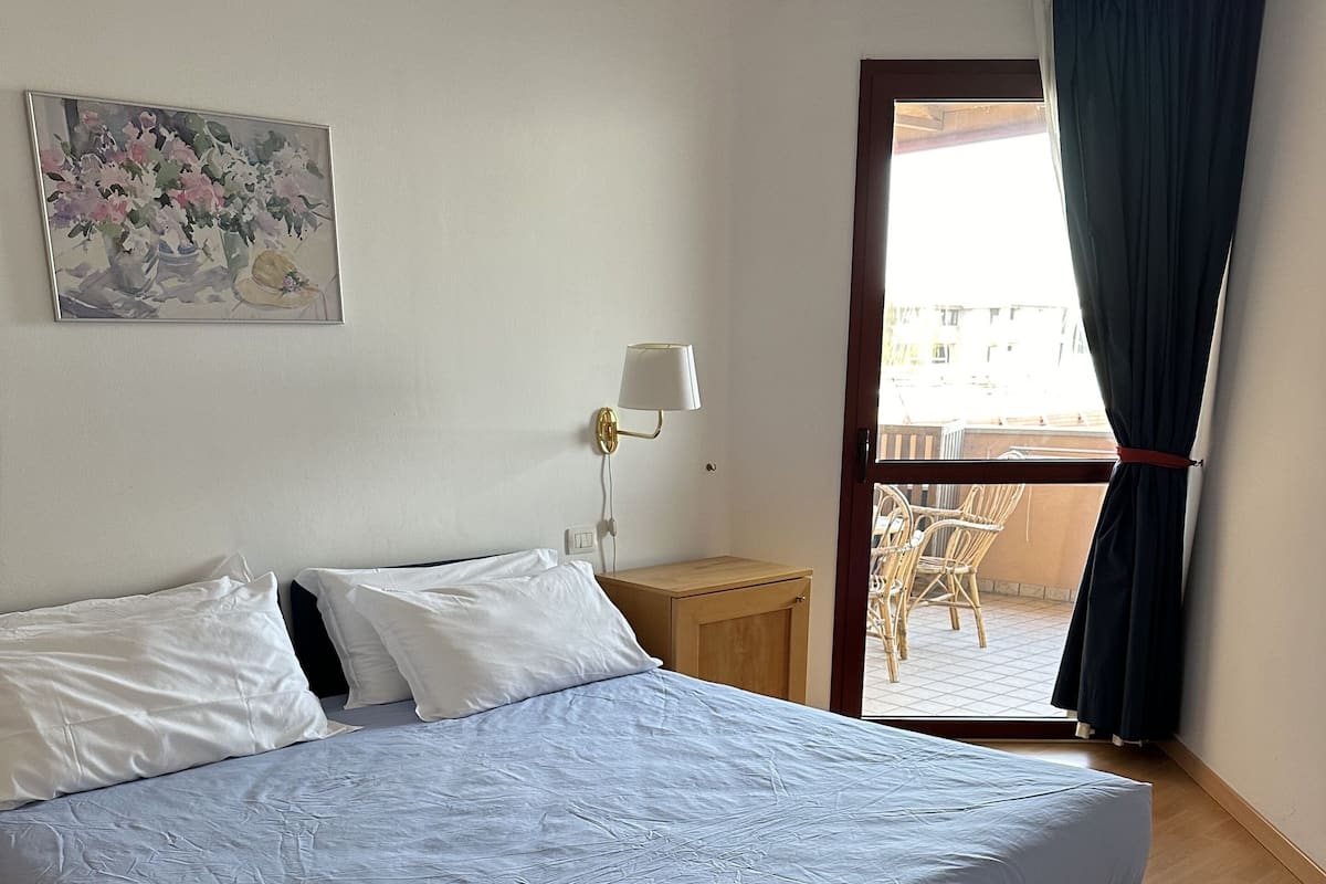 Une chambre à coucher comprenant un lit, une table de chevet, une lampe, un tableau et donnant sur le balcon extérieur.