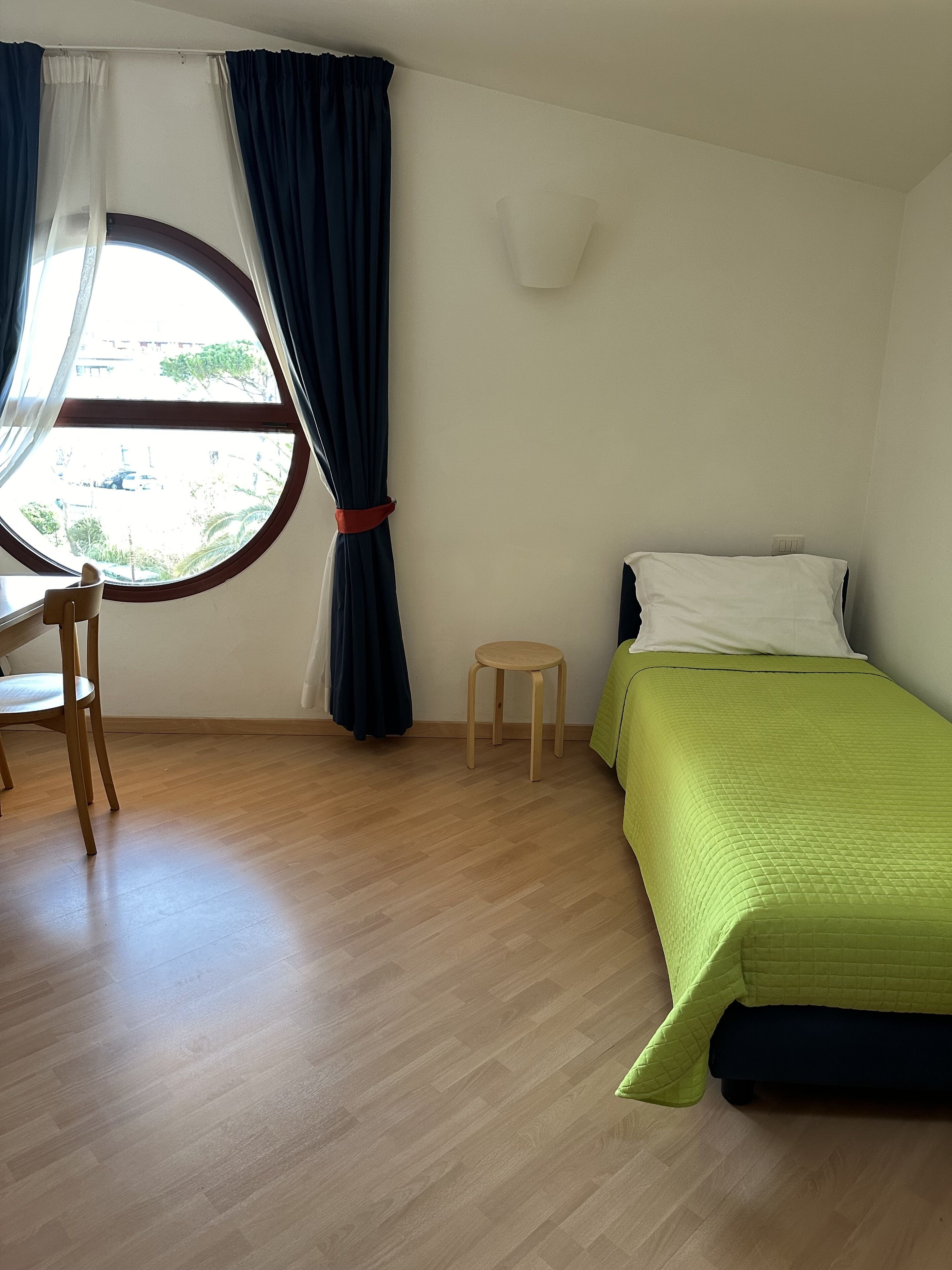 A room with a bed, a chair, a small stool, a window with curtains, and a wall-mounted light fixture.