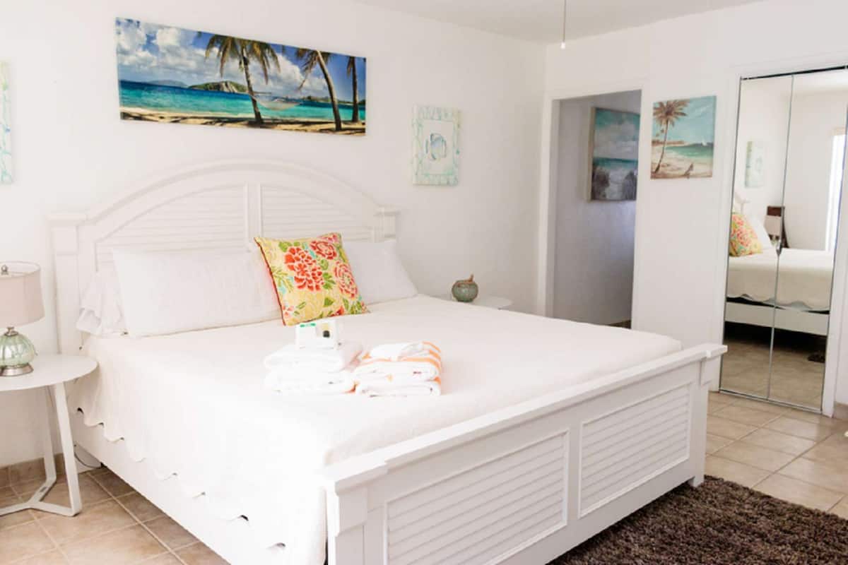 Un dormitorio con cama blanca, mesita de noche, lámpara y un cuadro con temática de playa en la pared.