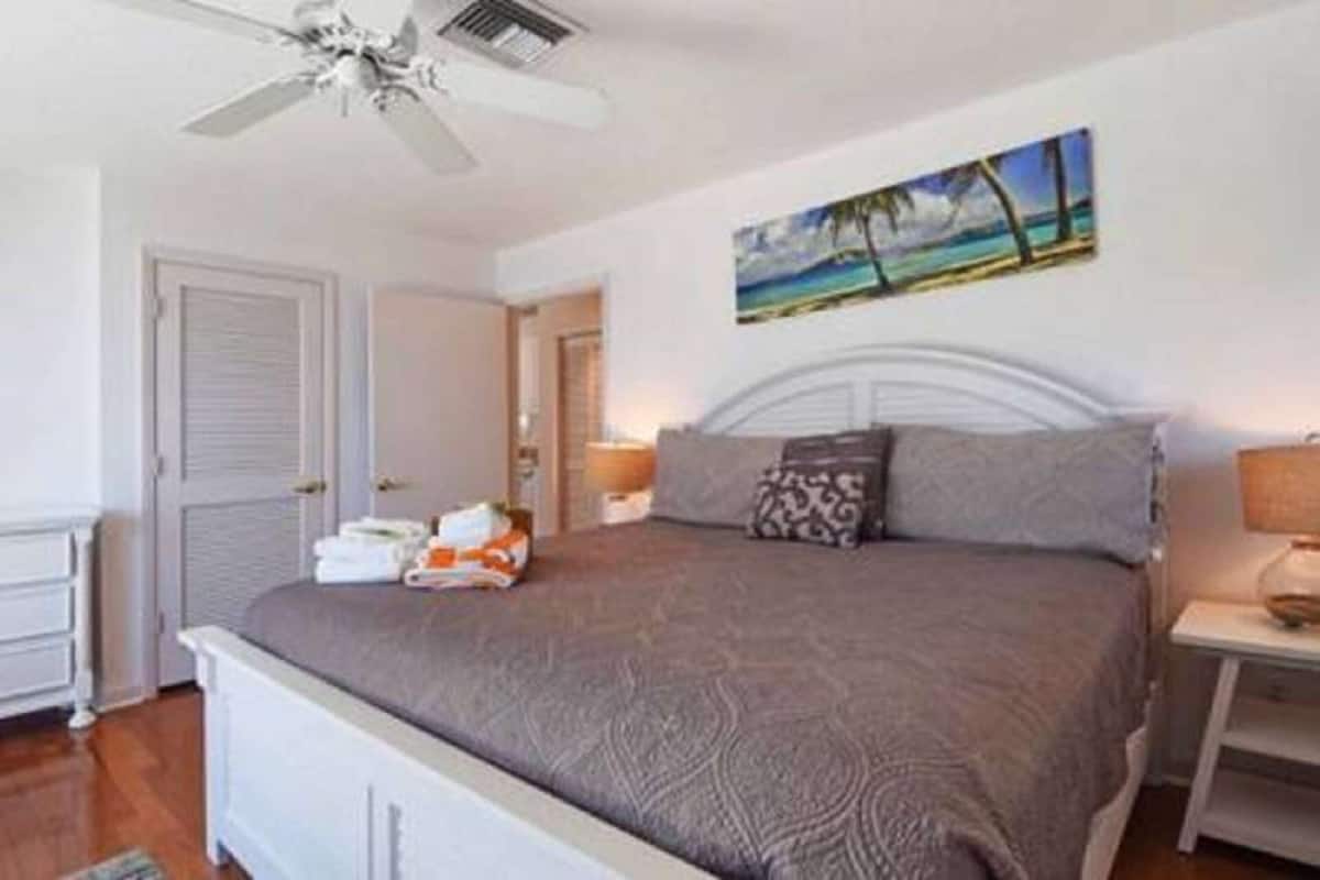 Un dormitorio con cama, ventilador de techo, un cuadro de una playa y una mesita de noche con lámpara.