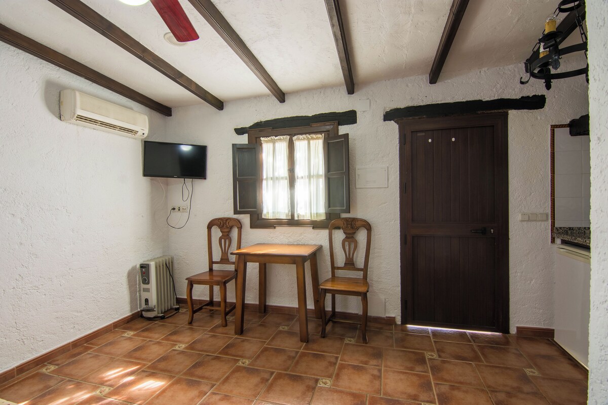 Quarto com piso de azulejos, uma mesa de madeira, duas cadeiras, televisão fixada na parede, uma janela com cortinas, uma porta e um aparelho de ar condicionado.