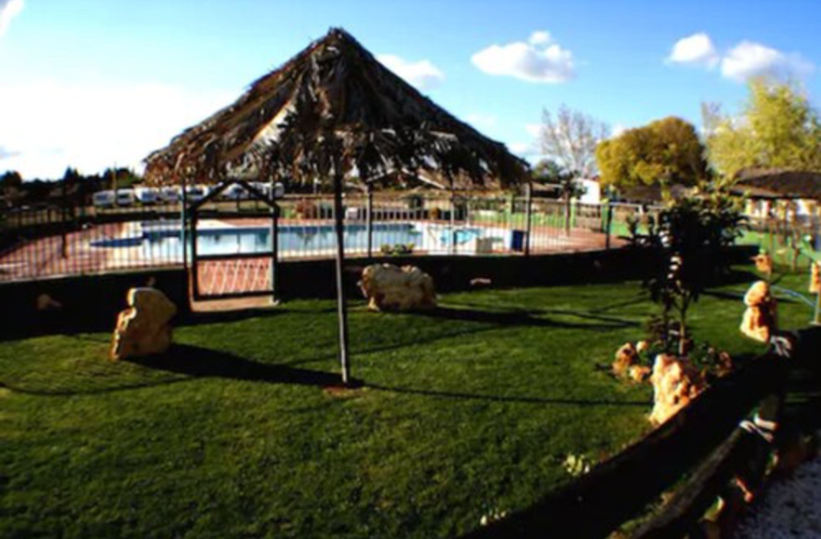 Un área de piscina con una estructura de techo de paja, piscina cercada y césped con rocas y plantas.