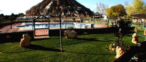 Un área de piscina con una estructura de techo de paja, piscina cercada y césped con rocas y plantas.