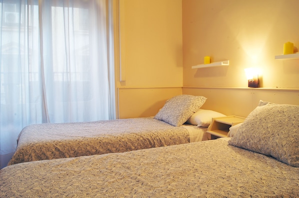 Hotellihuone, jossa on kaksi sänkyä, yöpöytä, pieni sivupöytä ja seinälle kiinnitetty hylly, jossa on kynttilöitä.