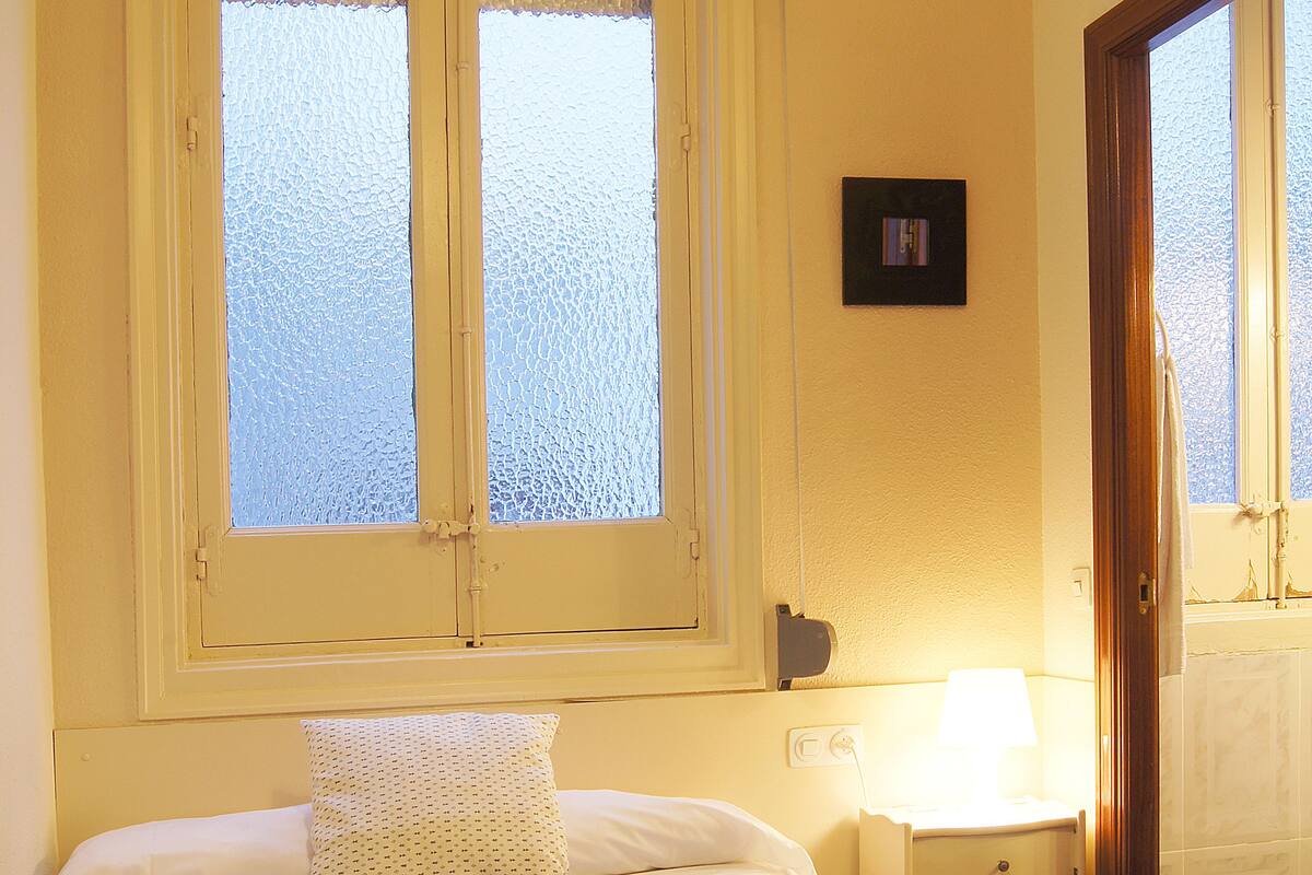 Une chambre à coucher avec un lit, une fenêtre à vitre dépolie, une table de chevet avec une lampe et une porte.