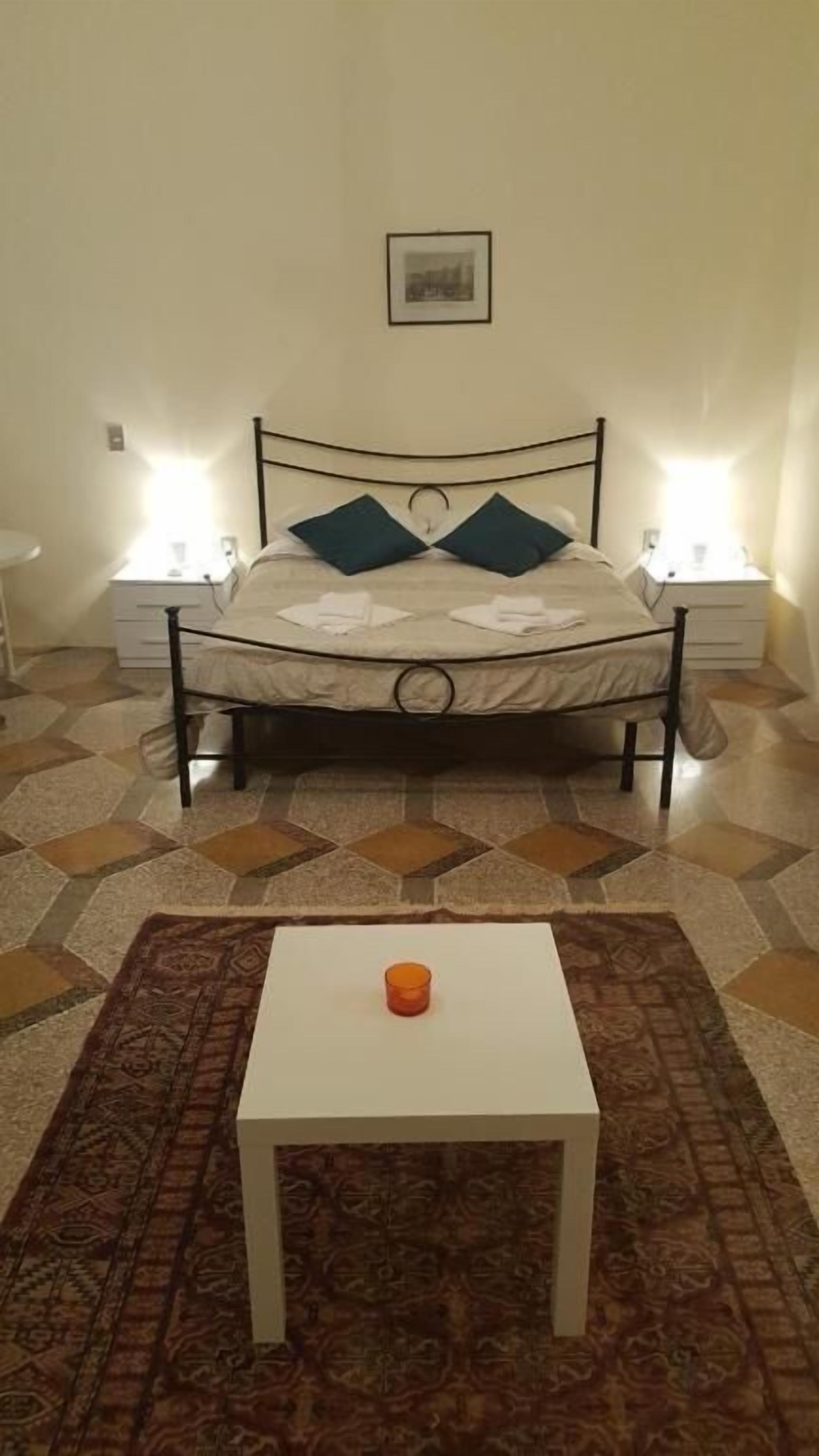 Uma cama com estrutura metálica, duas almofadas e um colchão branco. Há uma pequena mesa com um objeto laranja sobre ela. O quarto tem piso estampado e duas luminárias de cabeceira.