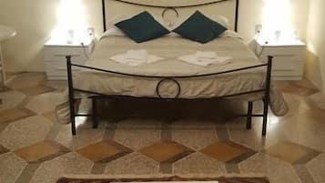 Uma cama com estrutura metálica, duas almofadas e um colchão branco. Há uma pequena mesa com um objeto laranja sobre ela. O quarto tem piso estampado e duas luminárias de cabeceira.