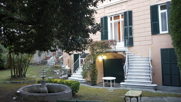 A building with a staircase leading to a balcony, a small garden with a fountain, and a door with a light above it. - Garden Suite (Genoa)
