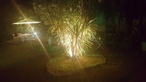 A well-lit outdoor area with a dining table and chairs, surrounded by lush greenery and a decorative tree. - Garden Suite (Genoa)