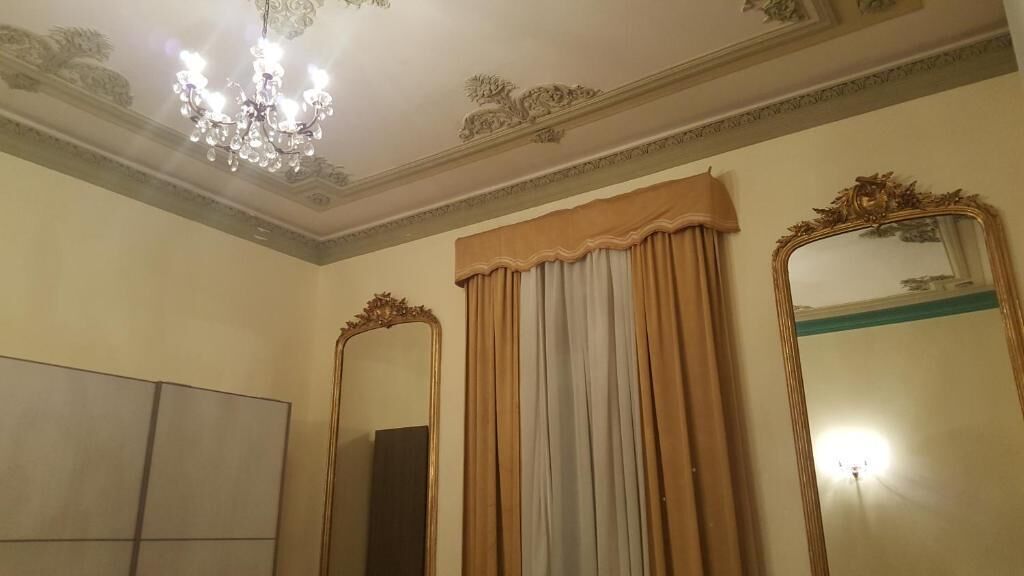Une pièce dotée d’un lustre, de deux miroirs aux cadres richement décorés et d’une grande fenêtre avec des rideaux.
