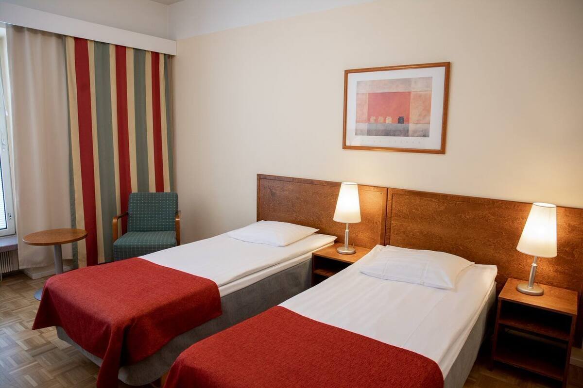 Une chambre d’hôtel avec deux lits simples, une chaise, une petite table et une fenêtre avec des rideaux à rayures.