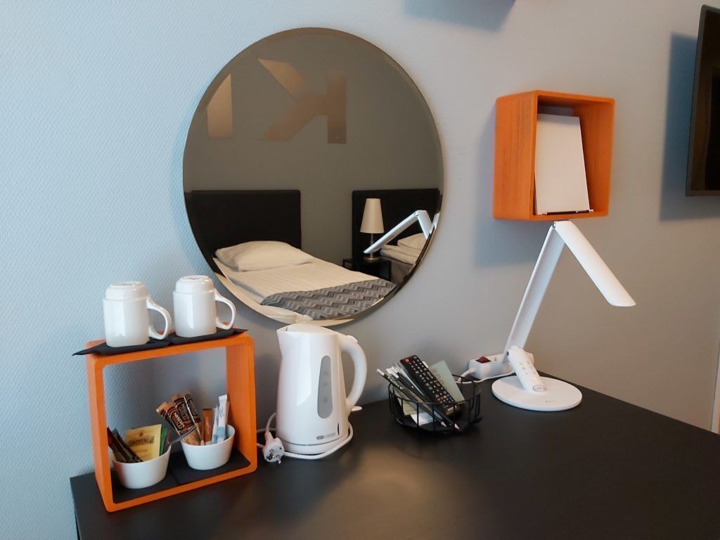 Ett hotellrum med en rund spegel, en säng med ett svart sänggavel, ett skrivbord med en lampa, en kaffebryggare och en hylla med muggar och en vattenkokare.