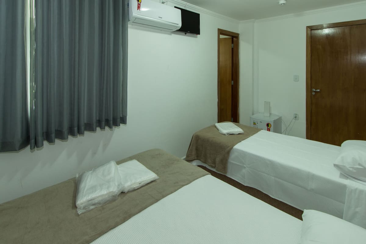 Una habitación de hotel con dos camas individuales, una ventana con cortinas, un equipo de aire acondicionado y una puerta de madera.