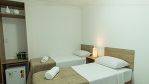 A hotel room with two single beds, a small refrigerator, a wooden shelf with decorative items, and a nightstand with a lamp. - Hotel Sabor e Fé (Aparecida)