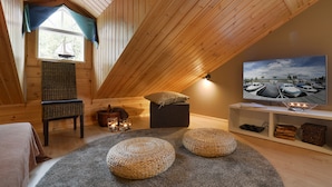 Acogedor dormitorio en ático con vigas de madera, televisor, mobiliario de mimbre y vistas a barcos por una ventana.
