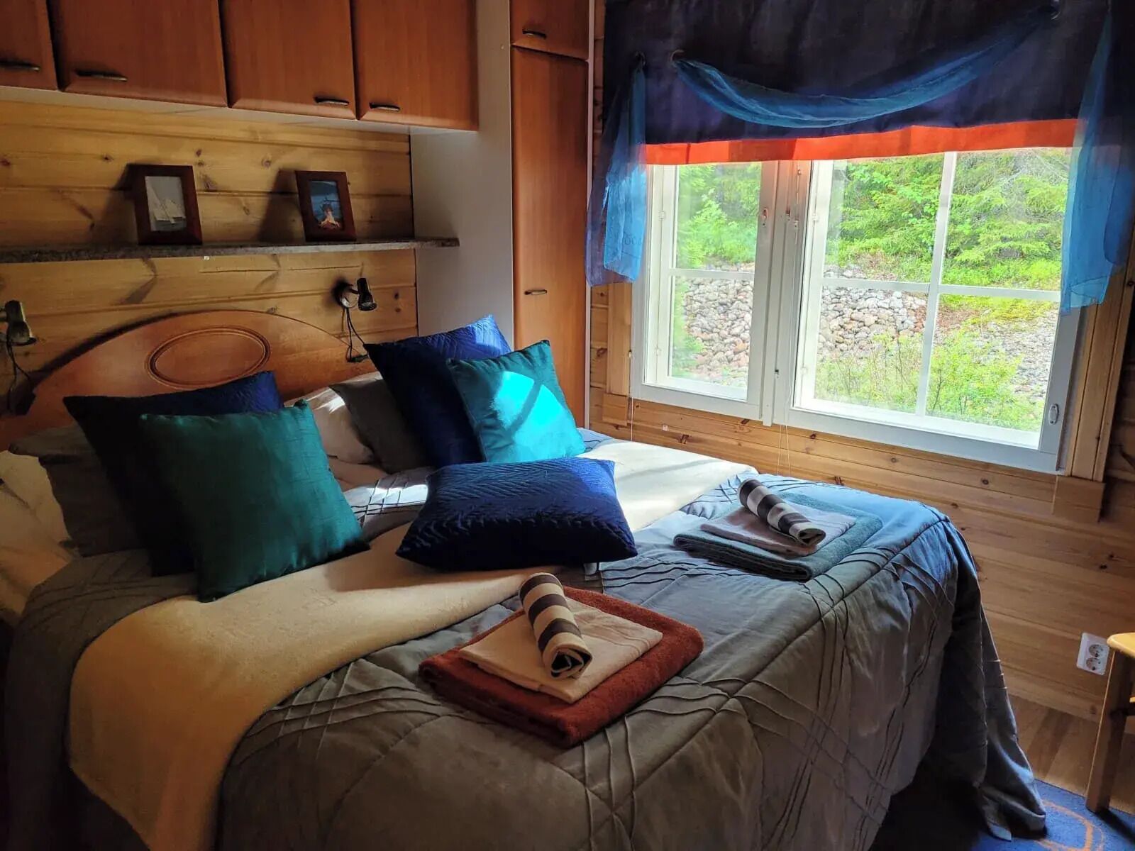 Un dormitorio de cabaña de madera con una cama, almohadas, una colcha y una ventana con vistas a la vegetación.