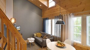 Una sala de estar acogedora con un sofá gris, una mesa de comedor con fruta, una escalera de madera y un ventanal con cortinas.