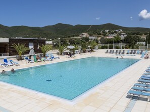 A rectangular swimming pool with clear blue water, surrounded by lounge chairs and umbrellas, set against a backdrop of hills and houses. - Sea-View holiday home in Northern Corsica (Belgodere)
