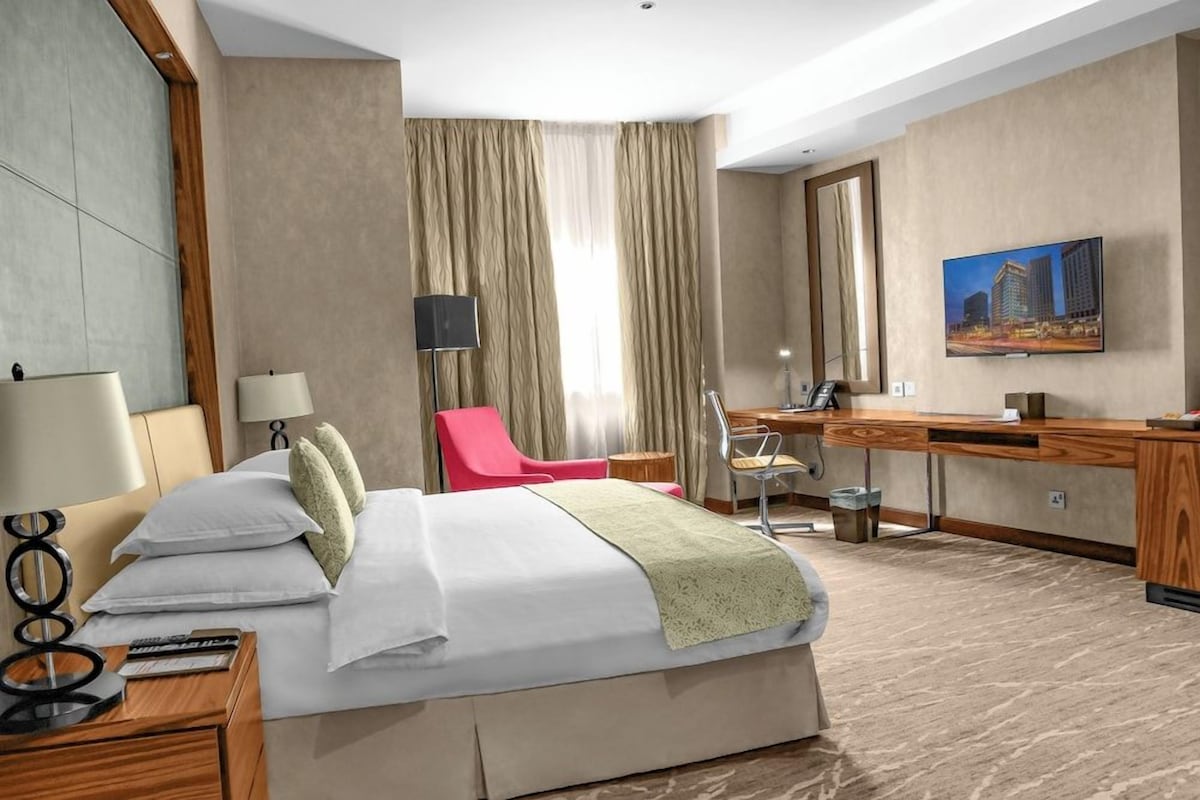 A hotel room with a bed, desk, chair, TV, and a painting on the wall.