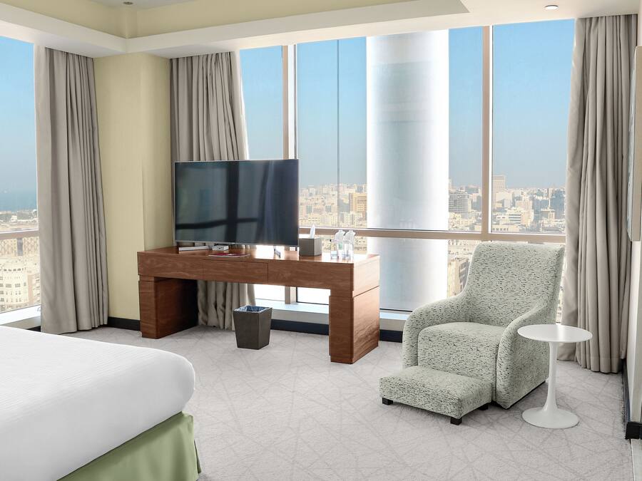 Quarto de hotel com cama, escrivaninha com televisão, cadeira e vista para a cidade.