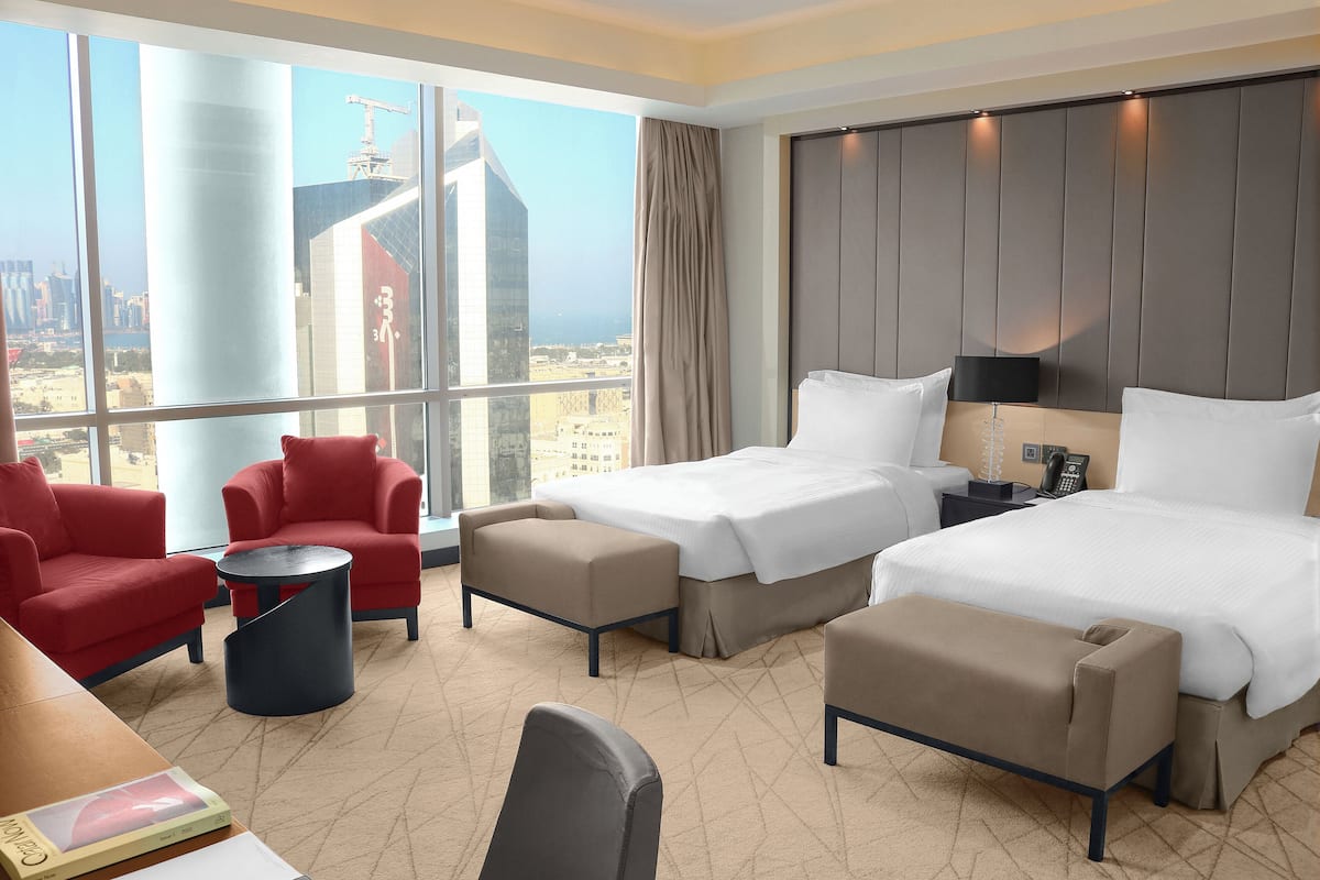A hotel room with two beds, red and beige armchairs, a small round table, and a view of a cityscape with tall buildings.