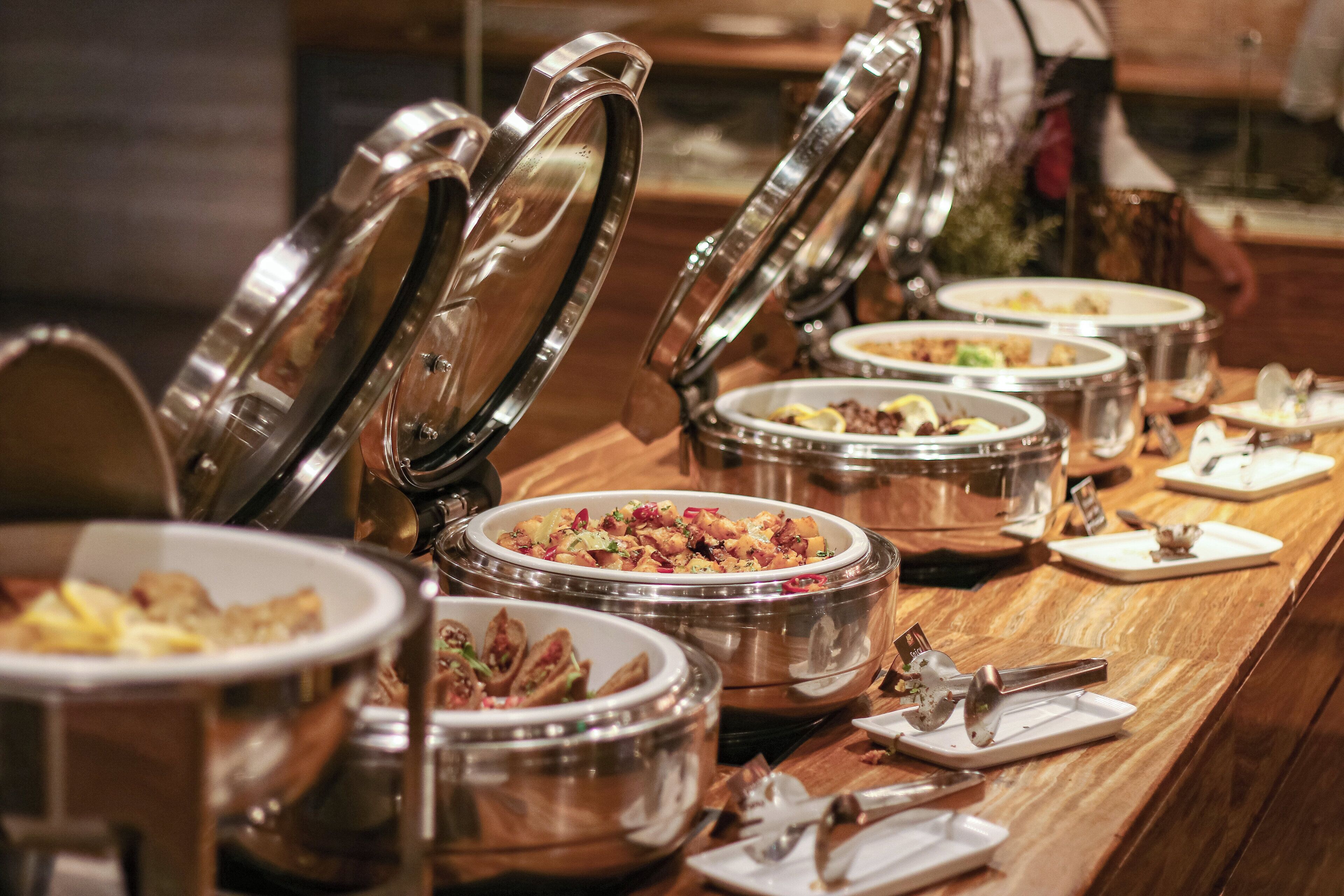 A buffet setup with multiple stainless steel chafing dishes on a wooden table, each with a lid.