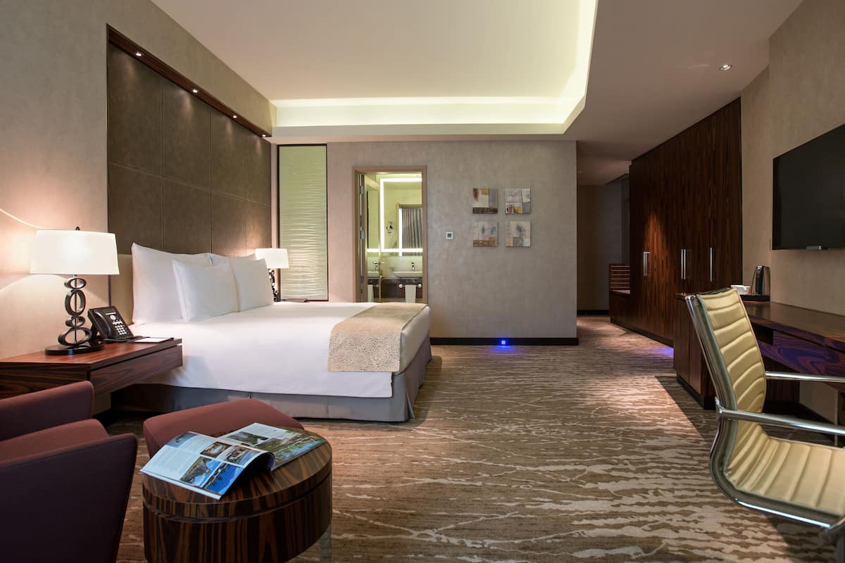 A hotel room with a bed, bedside lamps, a desk with a telephone, a chair, a round table with a magazine, a mirror, and a television on the wall.