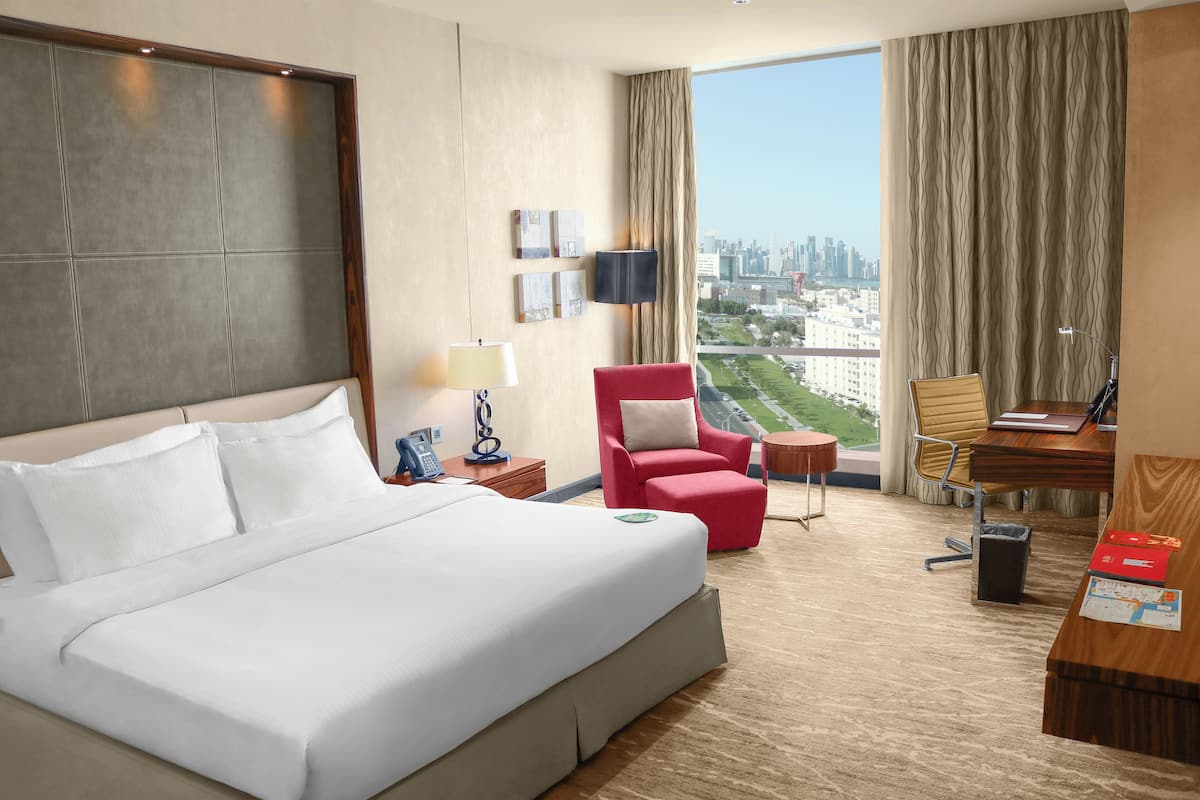 A hotel room with a large bed, a desk, a red armchair, and a view of the city.