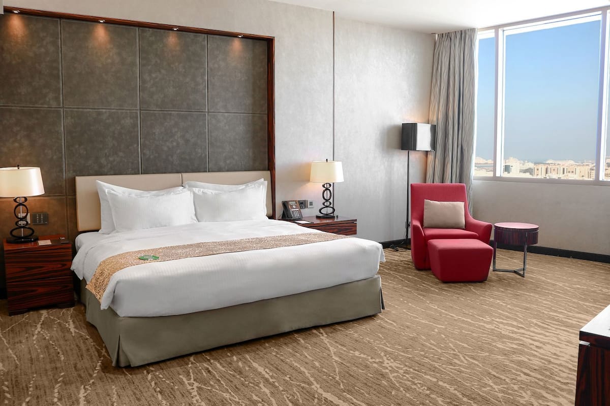 A hotel room with a large bed, two bedside tables with lamps, a red armchair, a small round table, and a view of the city through large windows.