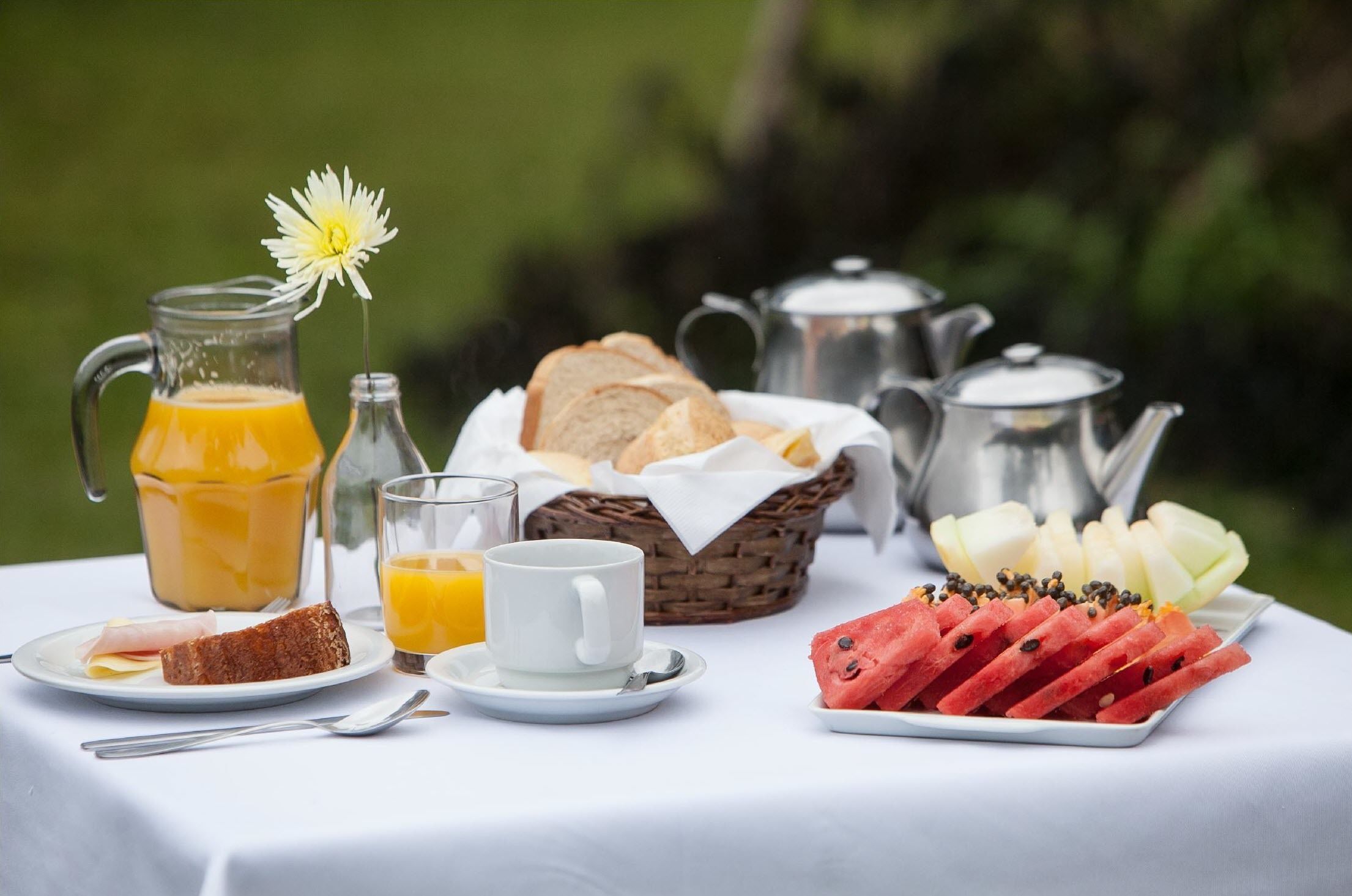 Morgenmad med en kande appelsinjuice, et glas appelsinjuice, en kop, en tallerken med ristet brød, en kurv med brød, en tallerken med vandmelon og en tepotte.