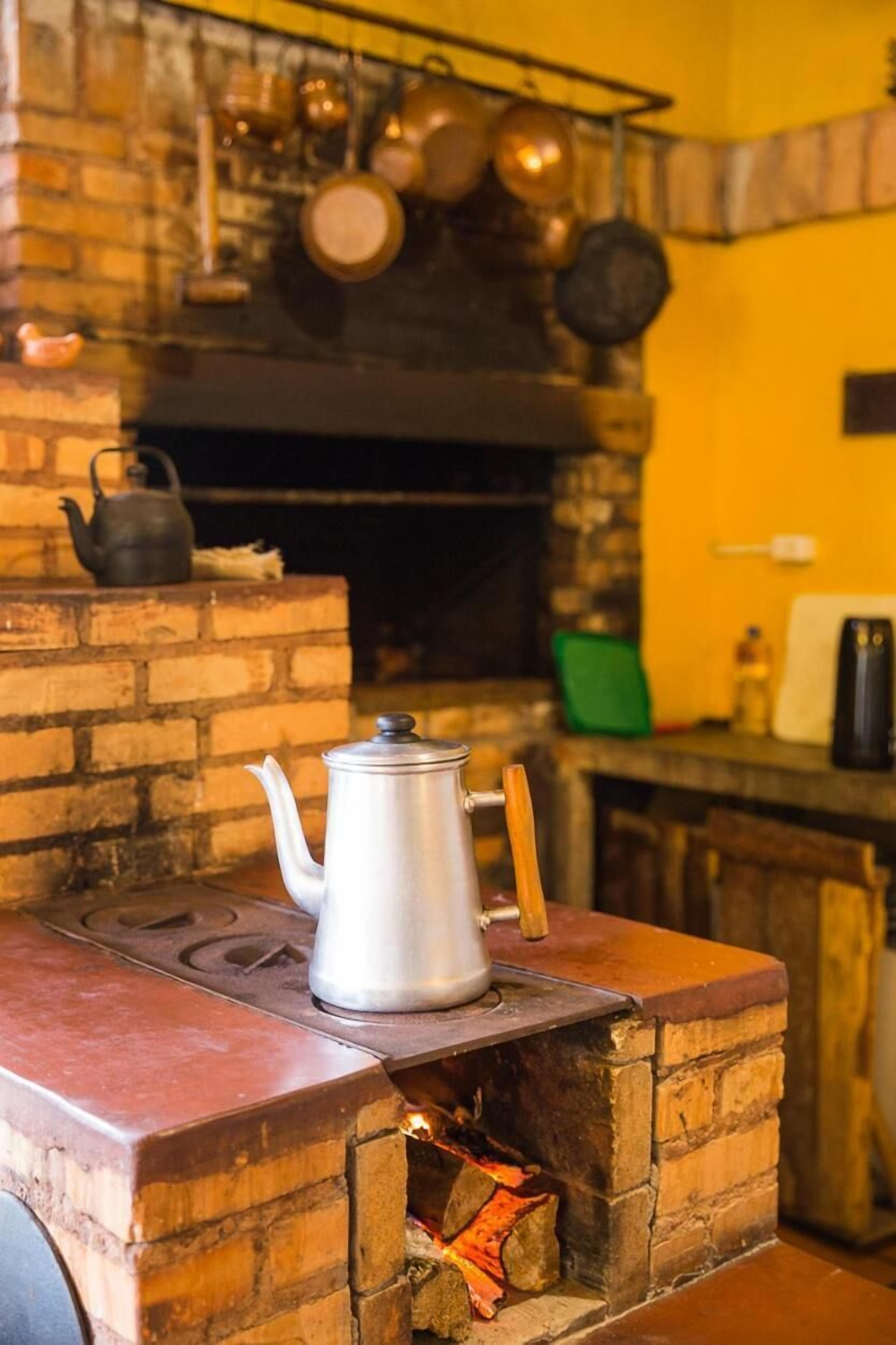 A brick oven with a kettle on top, a pot rack above, and a green chair in the background.